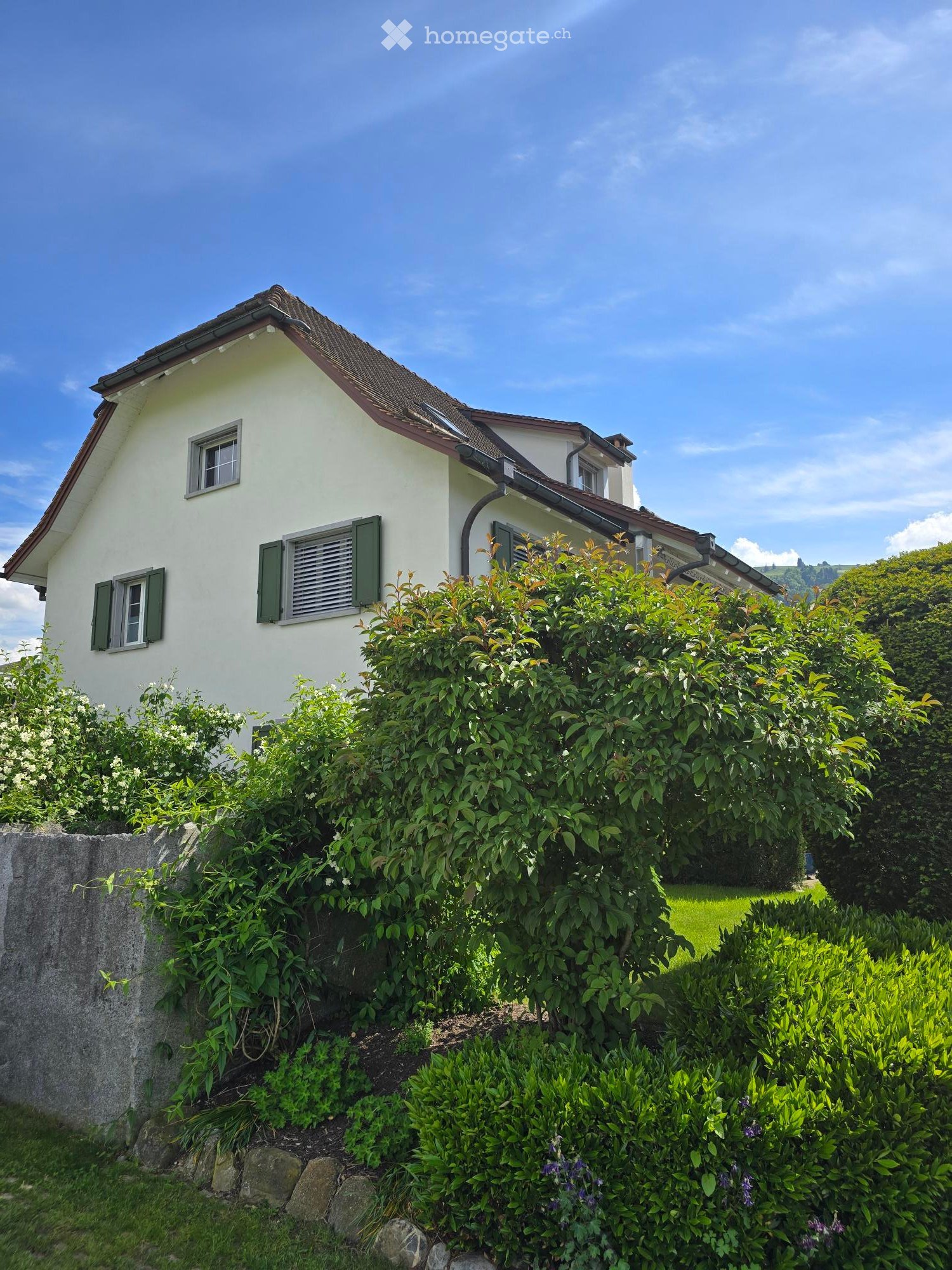 A two-story house with white walls, green shutters, and a red-tiled roof, surrounded by lush greenery and bushes.