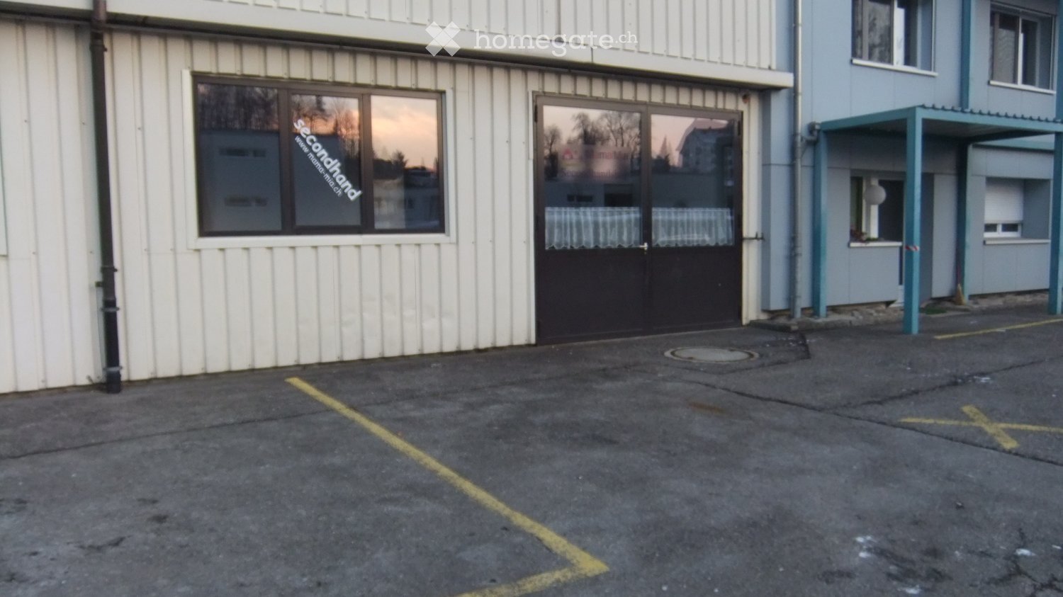 Exterior view of a multi-story building with white siding, large windows, and a covered entrance area. The ground is paved with marked parking spaces.