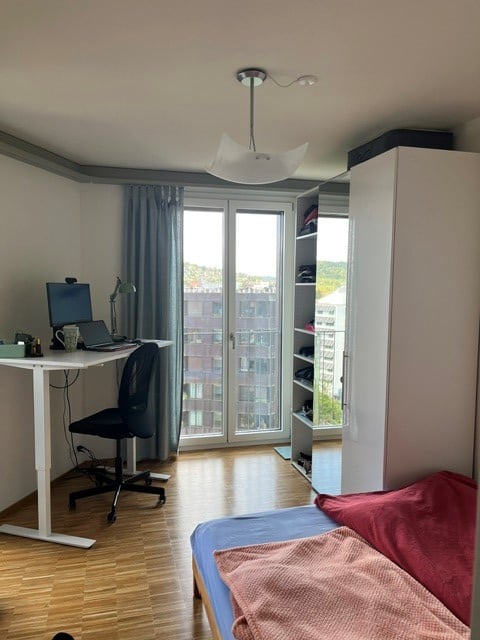 Modern, well-lit room with a bed, desk, chair, and laptop. Large window offers a view of the city.