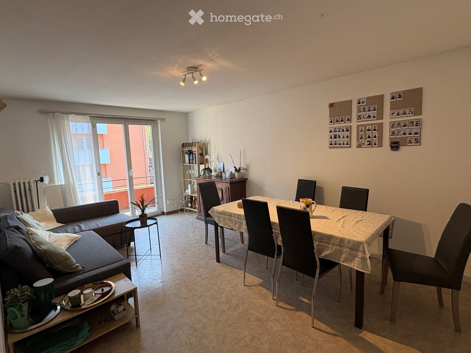 A modern living and dining area with a sofa, coffee table, dining table with chairs, and a bookshelf. The room features large windows with a sliding glass door leading to a balcony.