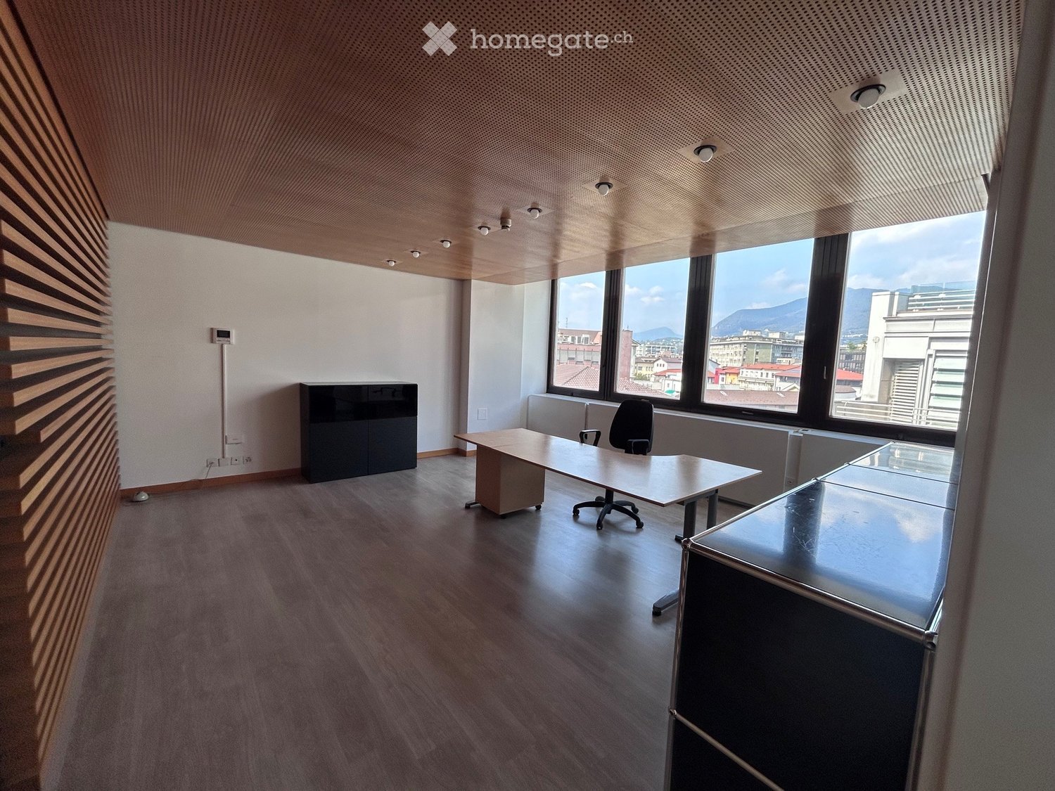 Modern office space with wooden flooring, large windows offering a cityscape view, a table, a chair, and a cabinet.