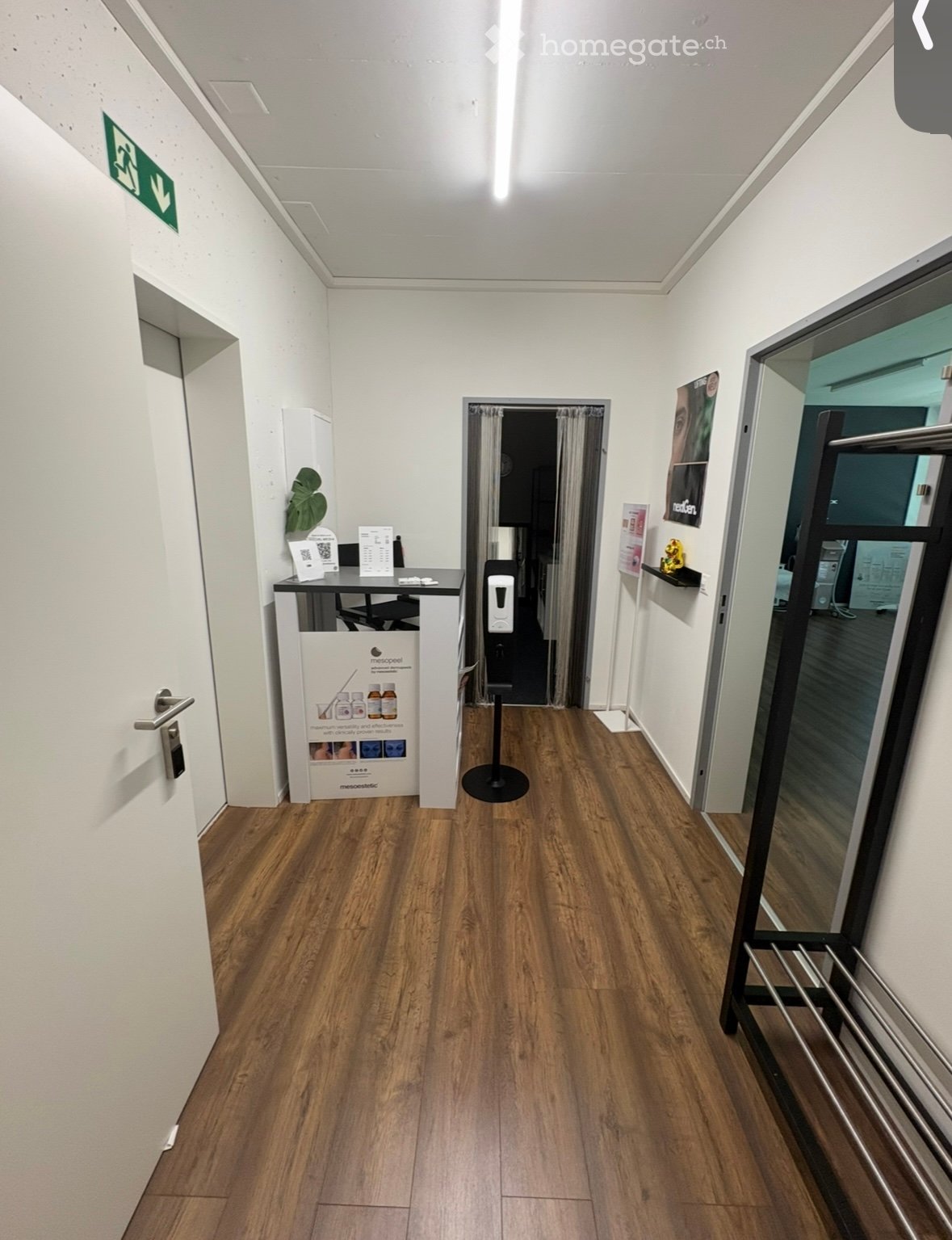 A clean, well-lit hallway with wooden flooring, white walls, and a reception area visible on the left side. There is a door with a silver handle, a small table with brochures, and a poster on the wall. A glass door with a metal frame is visible on the rig
