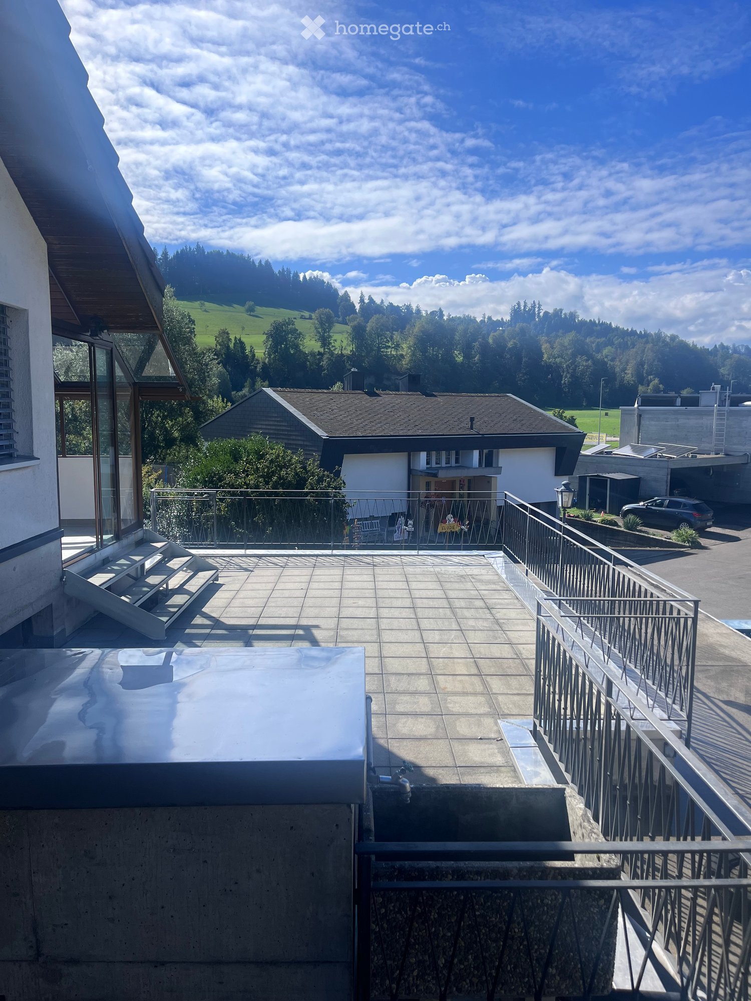 A modern rooftop terrace with a tiled floor, metal railing, and a scenic view of a green valley and houses.