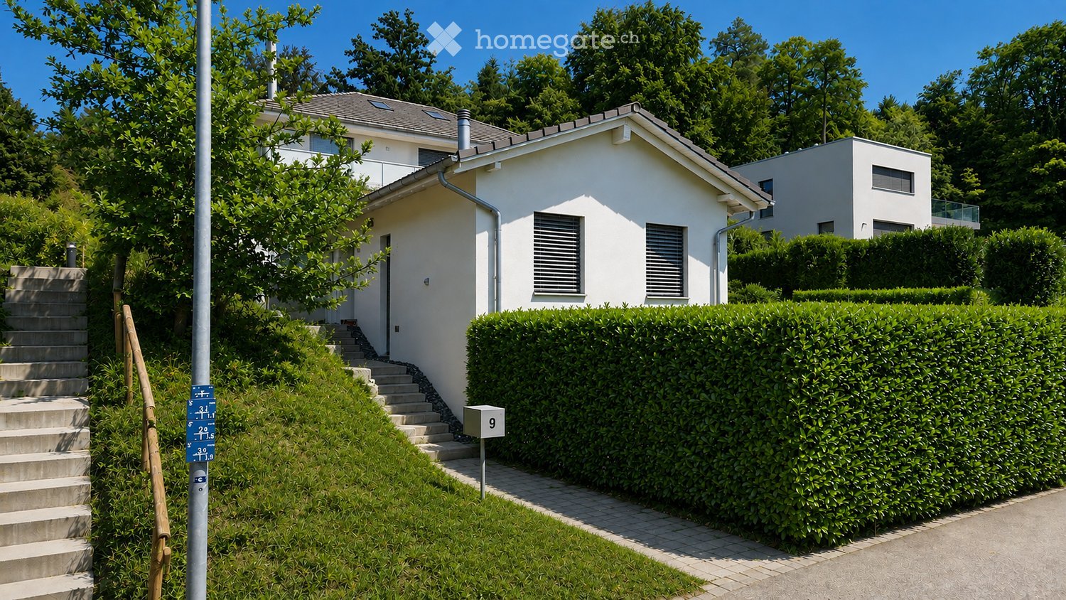 Single-story white house with a raised ground floor, surrounded by greenery and a neatly trimmed hedge. The entrance features concrete steps leading up to a door.