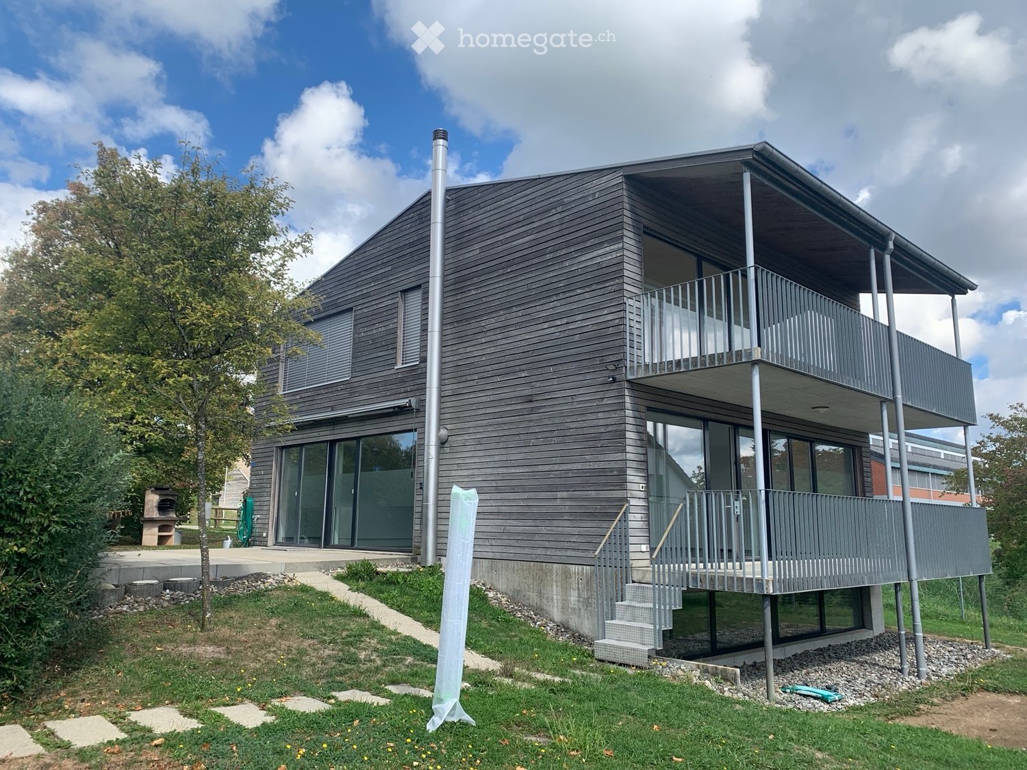 Modern two-story house with dark wooden exterior, large windows, and a balcony on the upper level. The house features a raised ground floor and is surrounded by a grassy area with a tree and a small pathway leading to the entrance.