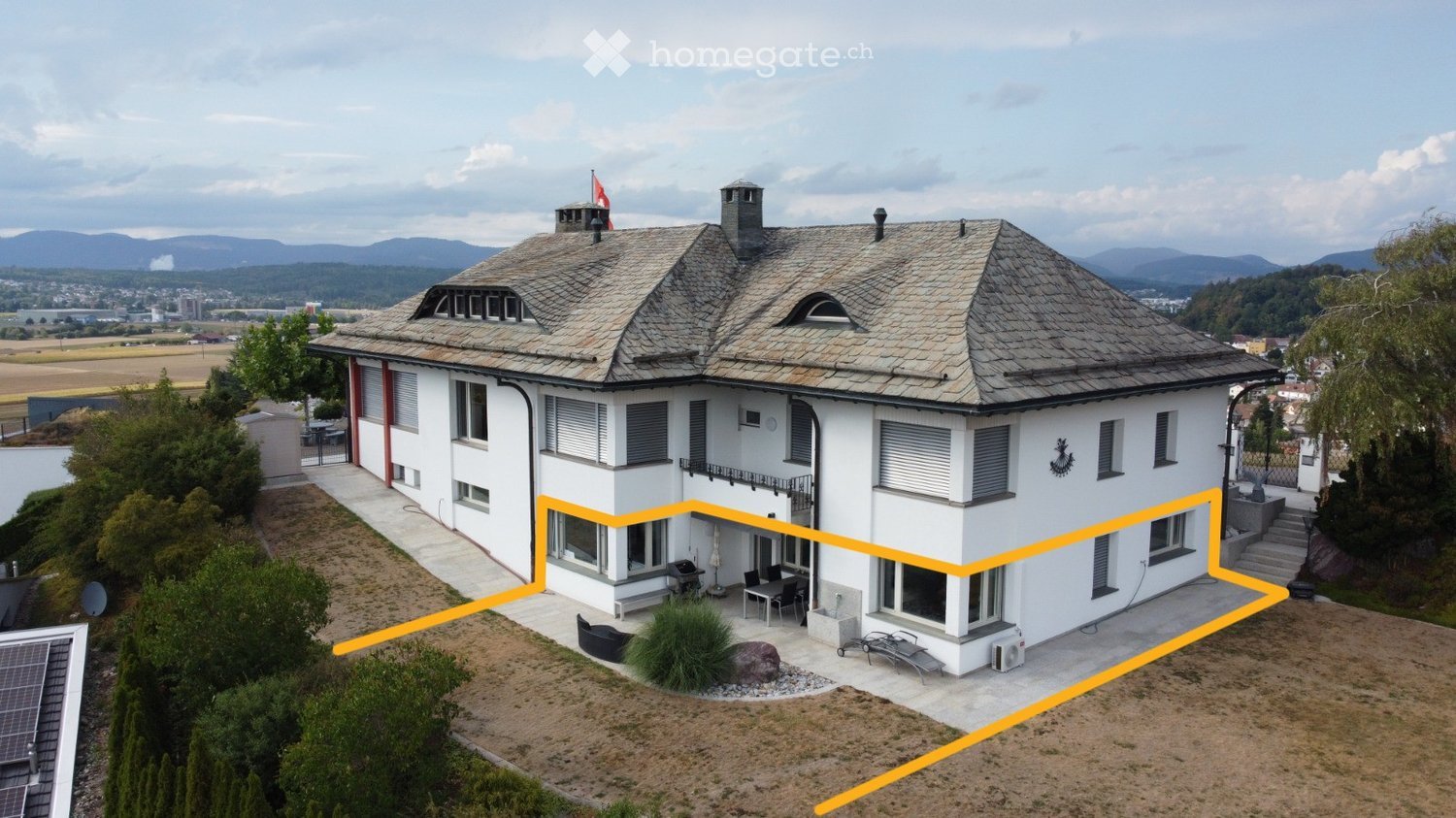 A two-story white house with a gray tiled roof, multiple chimneys, and a balcony. The house features large windows, shutters, and a small garden area with outdoor furniture.