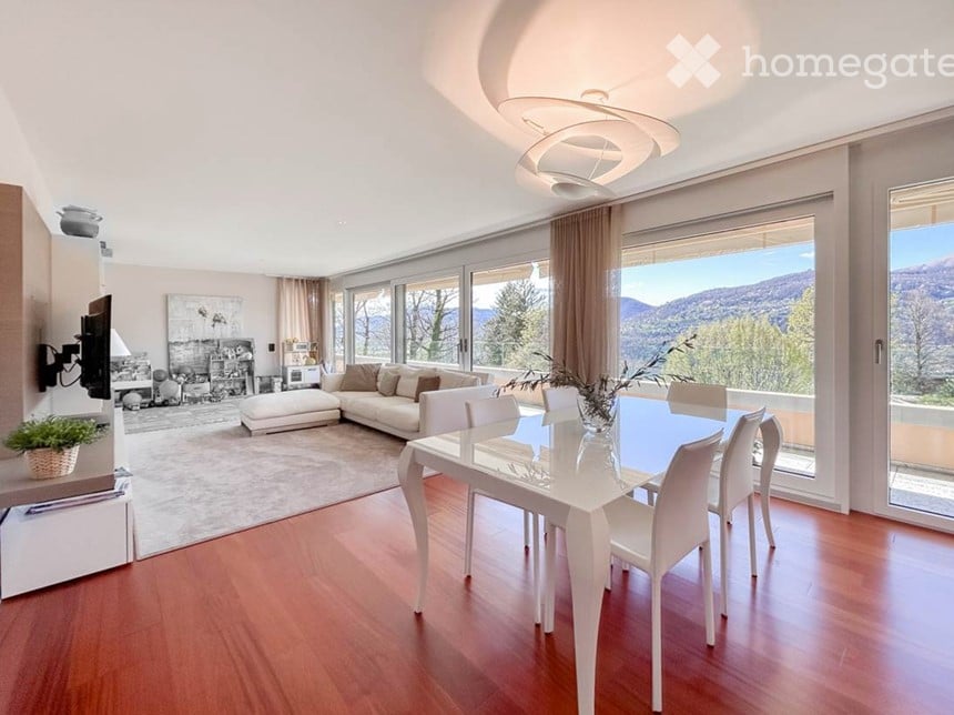 Bright, open-plan living and dining area with large windows offering a scenic mountain view. Features a white sectional sofa, a glass dining table with white chairs, a floor lamp, and a wall-mounted TV.