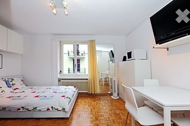 Cozy studio apartment with a bed, small table and chairs, refrigerator, microwave, fan, and TV. The room has wooden flooring and a window with a view of the balcony.
