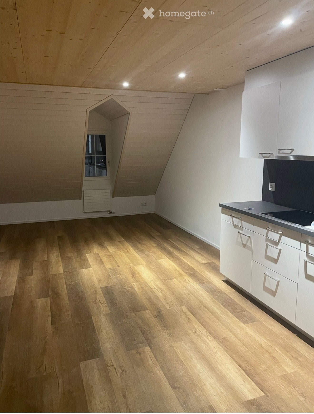 An unfurnished attic room with wooden flooring, white walls, and a small window. The room features built-in white cabinets and a countertop on the right side.