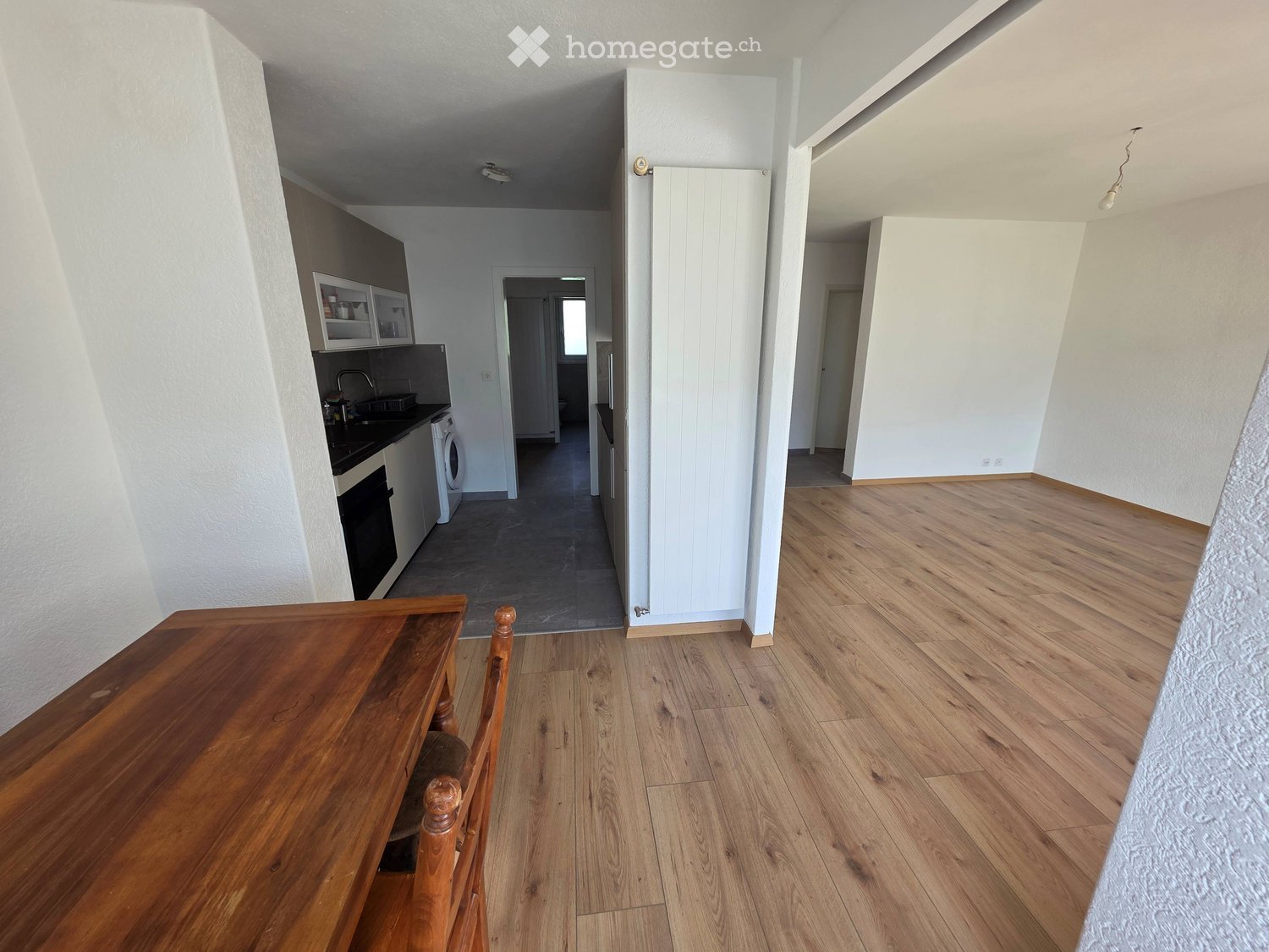 A furnished room with wooden flooring, a wooden table and chair, a kitchen area with cabinets, a sink, a faucet, a fridge, and an oven.
