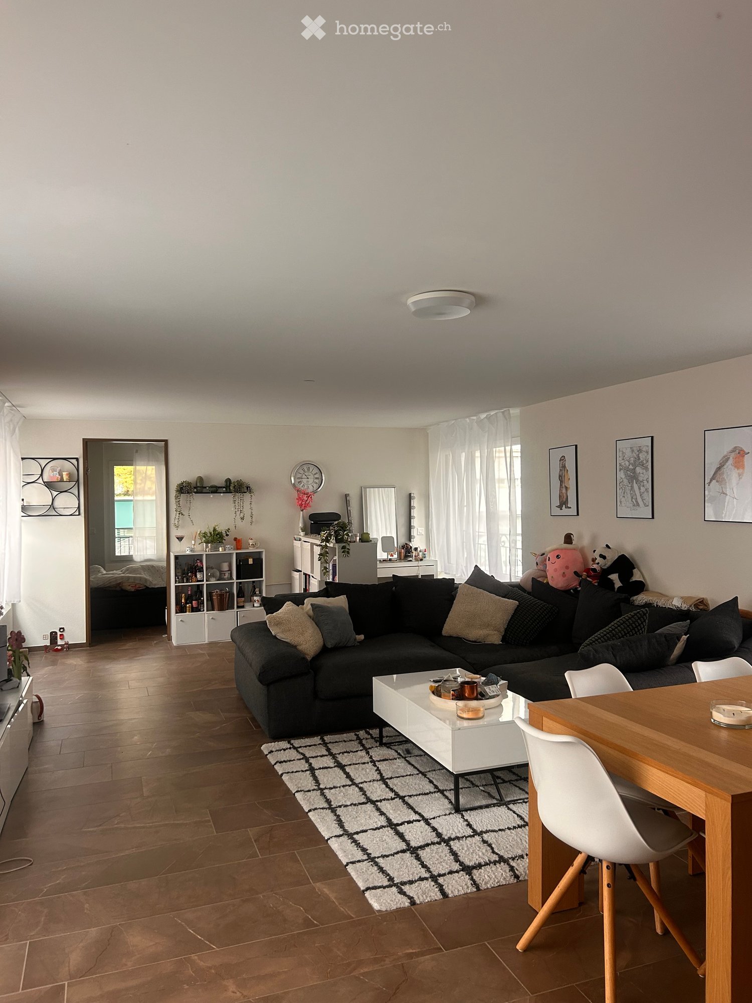 Modern living room with a black sectional sofa, white coffee table, dining table with chairs, and a shelf with decorative items. The room features wooden flooring, a patterned rug, and large windows with white curtains.