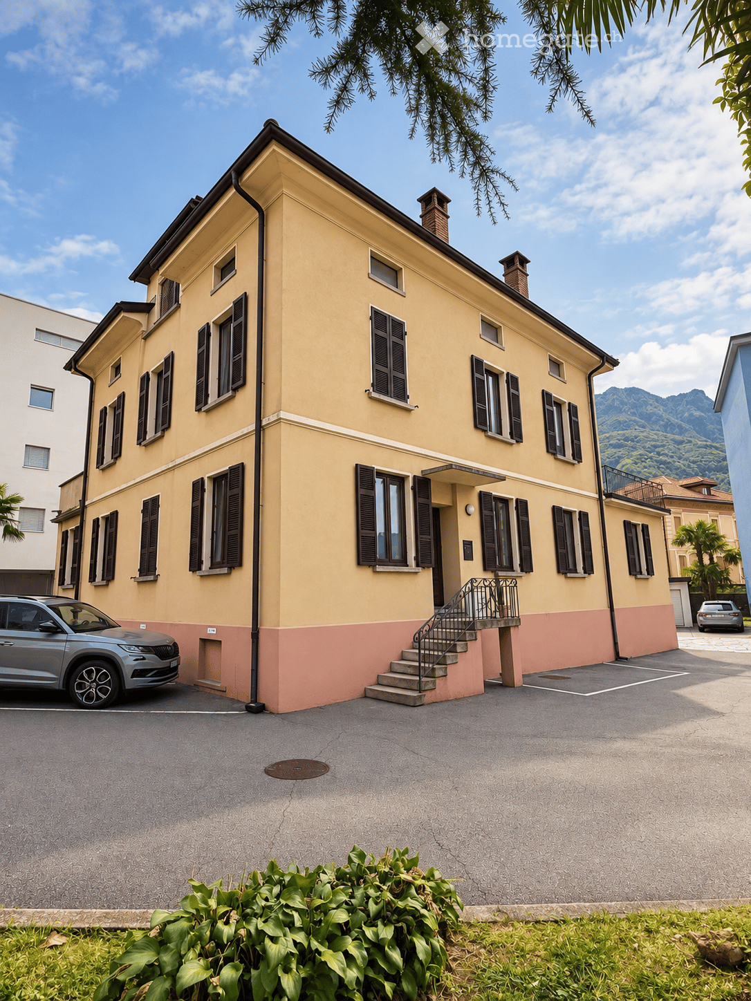 Two-story yellow house with black shutters, a small staircase leading to the entrance, and a parked car in front.