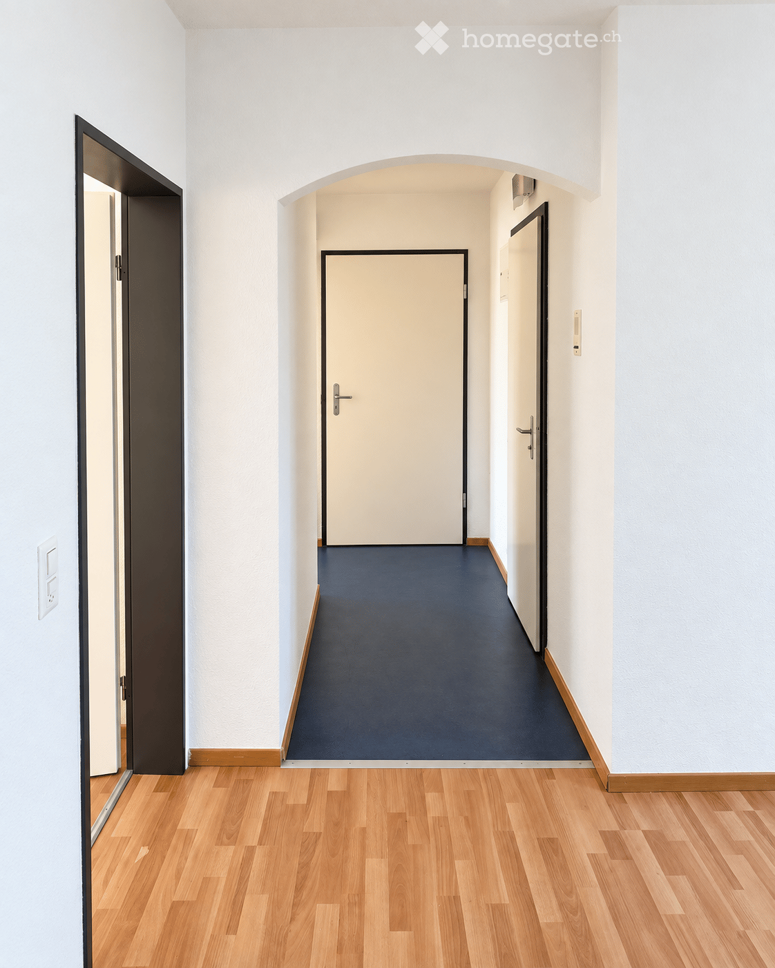 A modern hallway with light wooden flooring, white walls, and a dark blue carpeted section leading to a closed door.