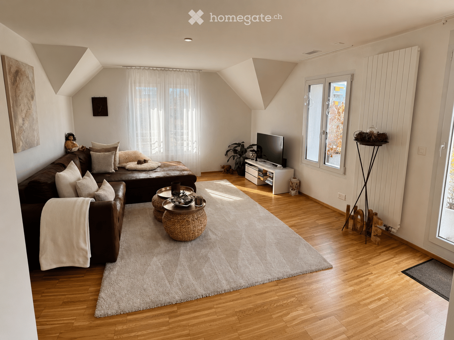 Modern living room with wooden flooring, large window with white curtains, brown leather sofa, coffee table, TV, and decorative items.