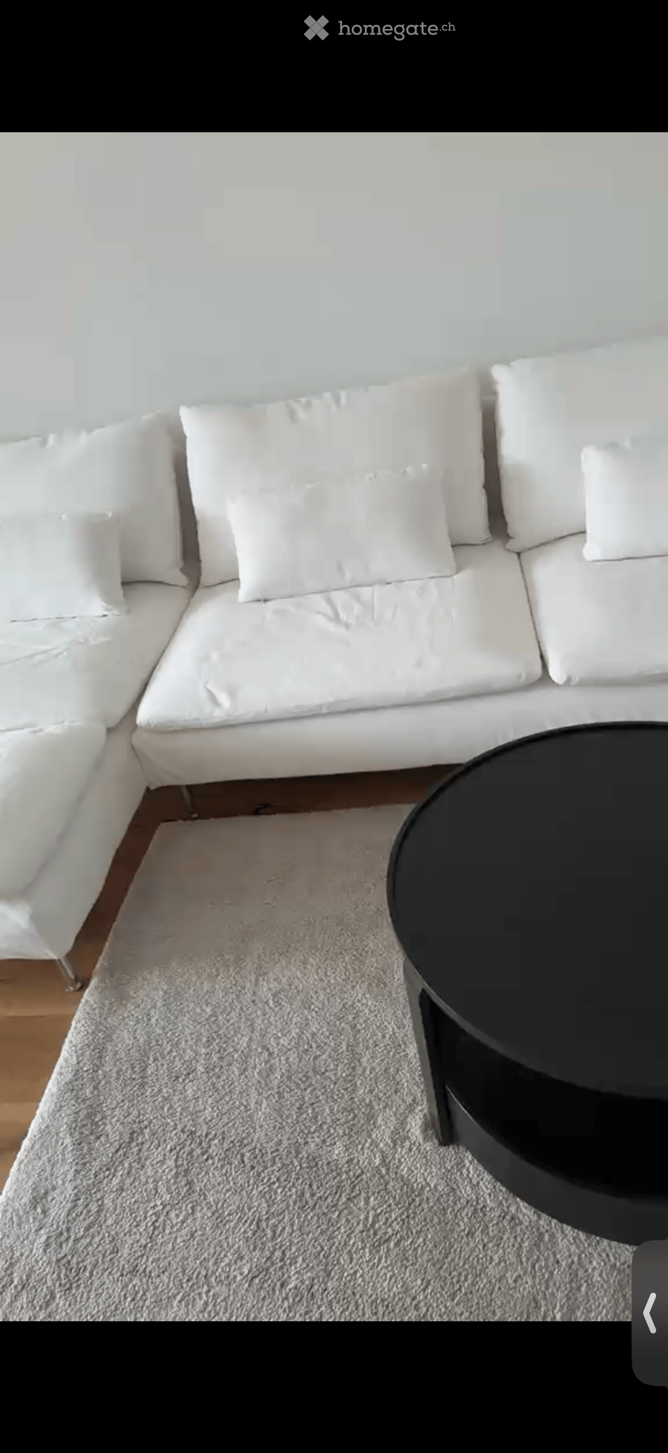 A modern living room with a white sectional sofa, multiple white cushions, a grey shag rug, and a black round coffee table.