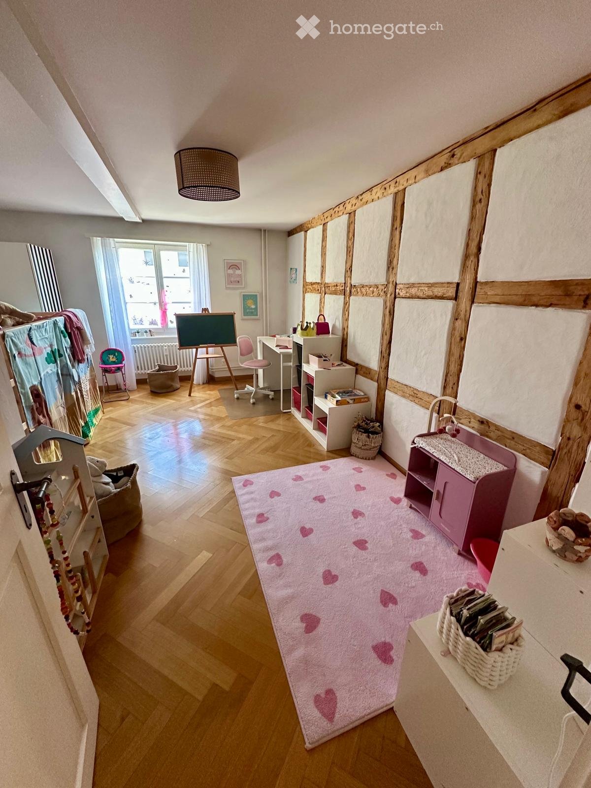 A cozy, well-lit room with wooden flooring, a pink heart-patterned rug, a small table with a chair, a cabinet, and various toys and items scattered around.