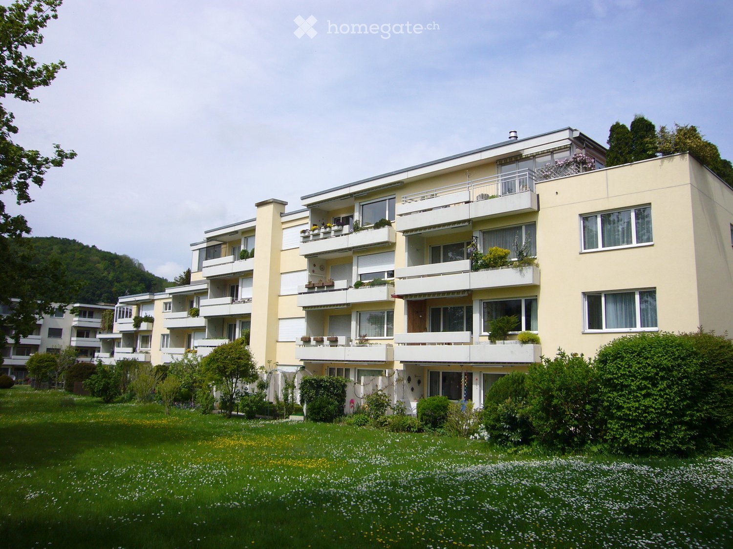 Multi-story residential building with balconies, large windows, and surrounding greenery.