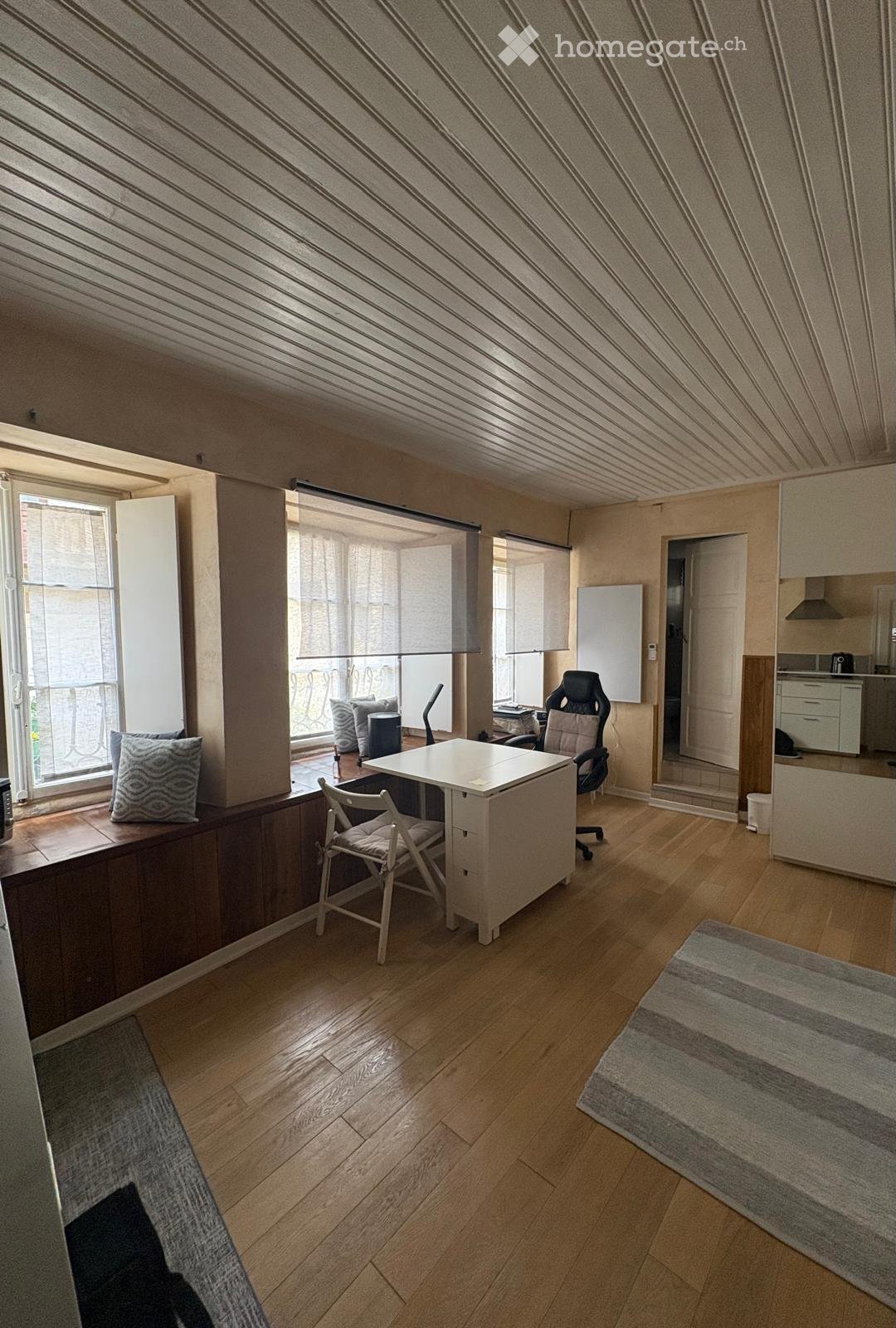Bright, modern room with wooden flooring, large windows, a desk with chairs, a kitchen area, and a balcony view.