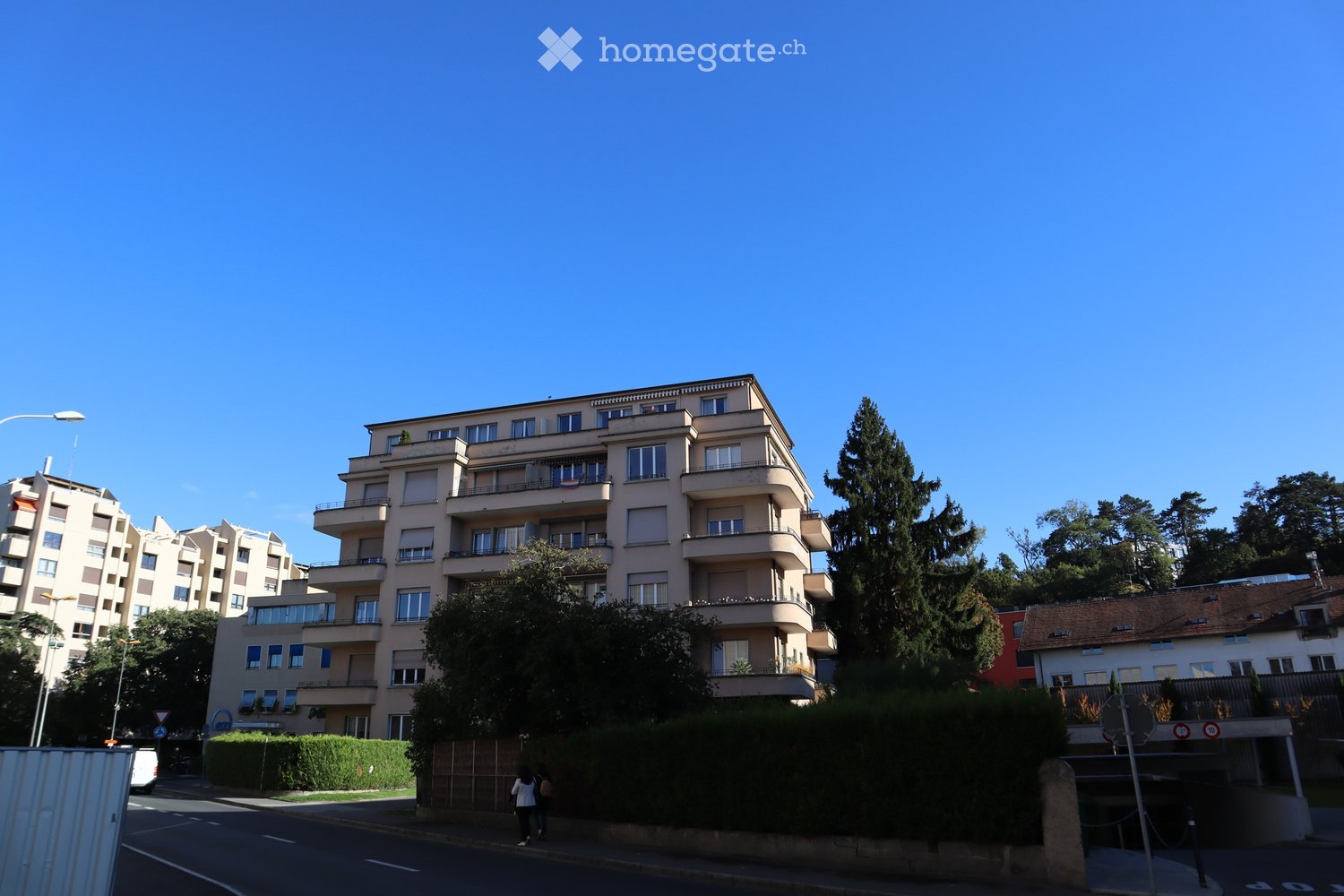 Multi-story residential building with balconies and large windows, surrounded by trees and greenery.