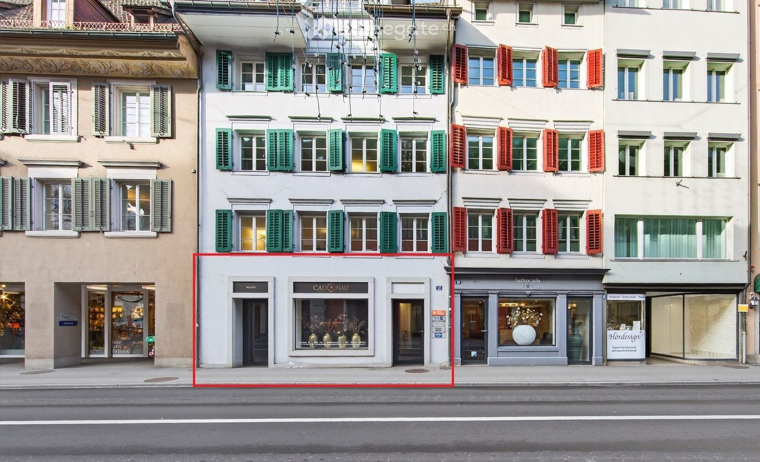A multi-story building with classic European architecture, featuring green and red shutters, large windows, and storefronts including 'CADONAU' and 'Gallerie Sofia'.