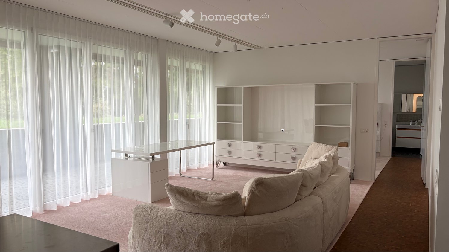 Modern living room with a light pink carpet, white walls, large windows with sheer curtains, a beige sofa, a glass coffee table, and a white shelving unit.