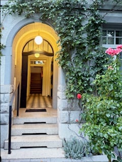 Entrance to a building featuring an arched doorway with a yellow-lit interior, flanked by greenery and flowers. The steps lead up to a wooden door with a small welcome mat.