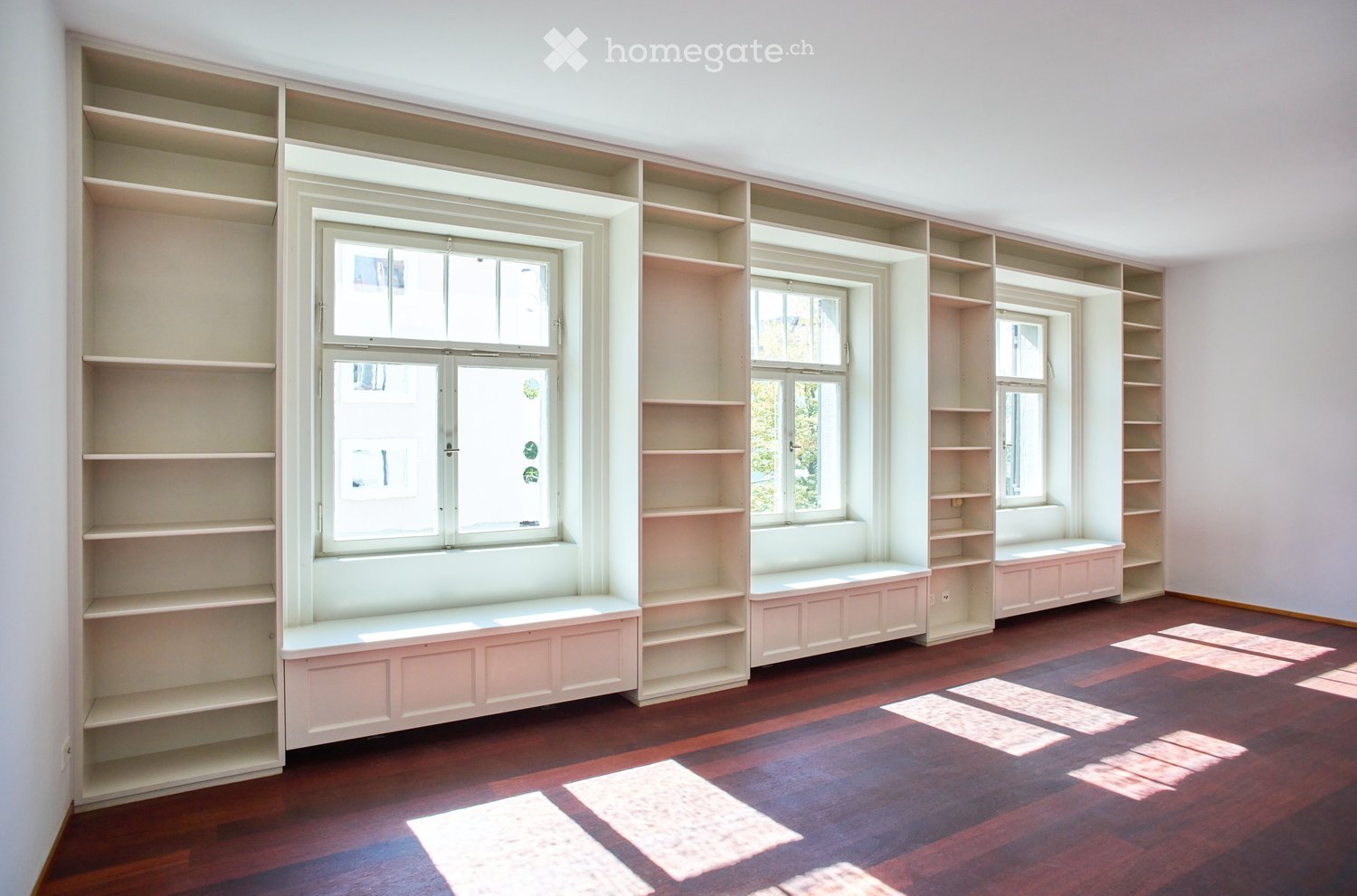 Empty room with built-in shelves and window seats, wooden flooring, and natural light from multiple windows.