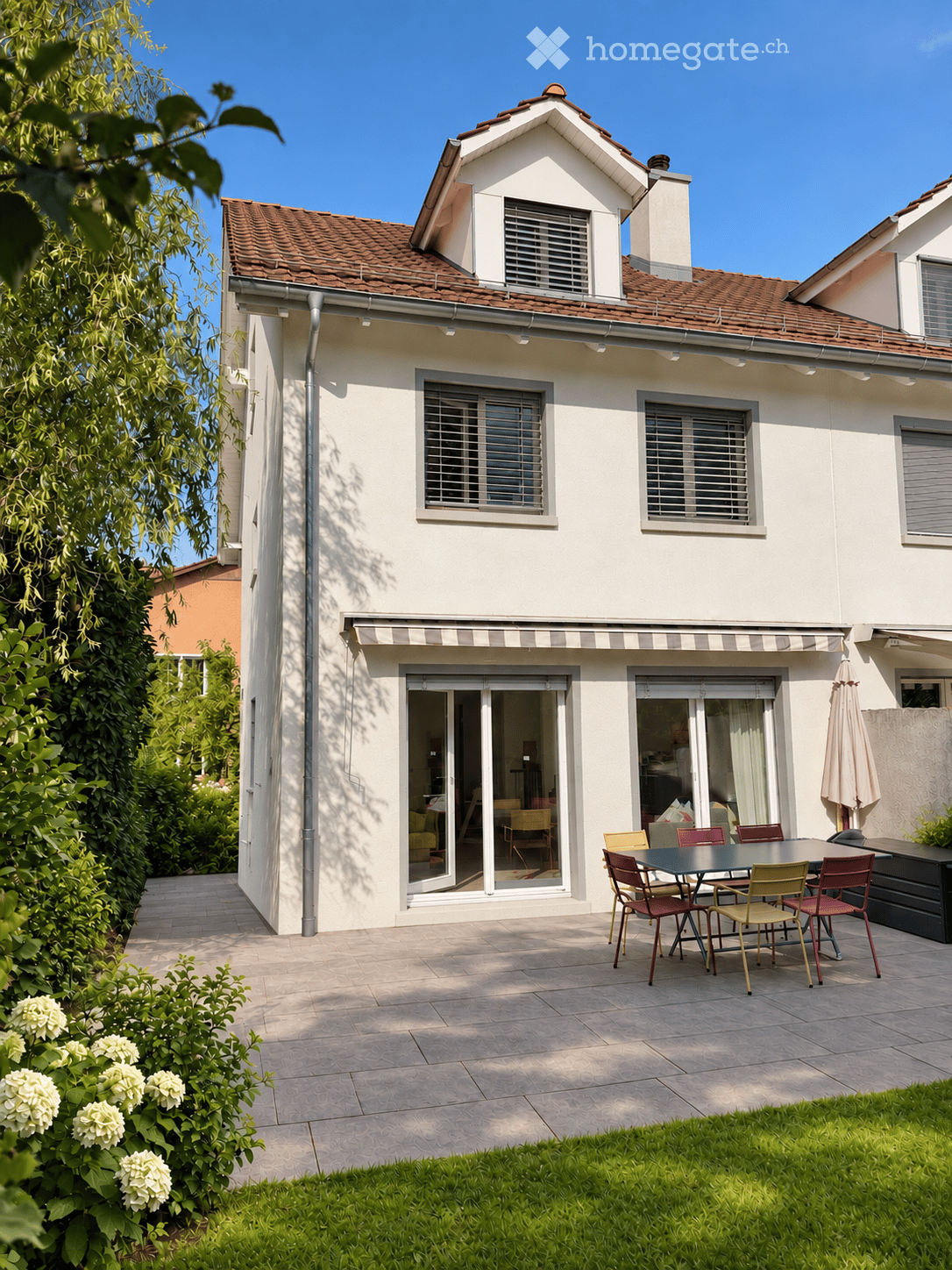 Two-story house with a tiled roof, white exterior, large windows, and a covered patio area with outdoor furniture.