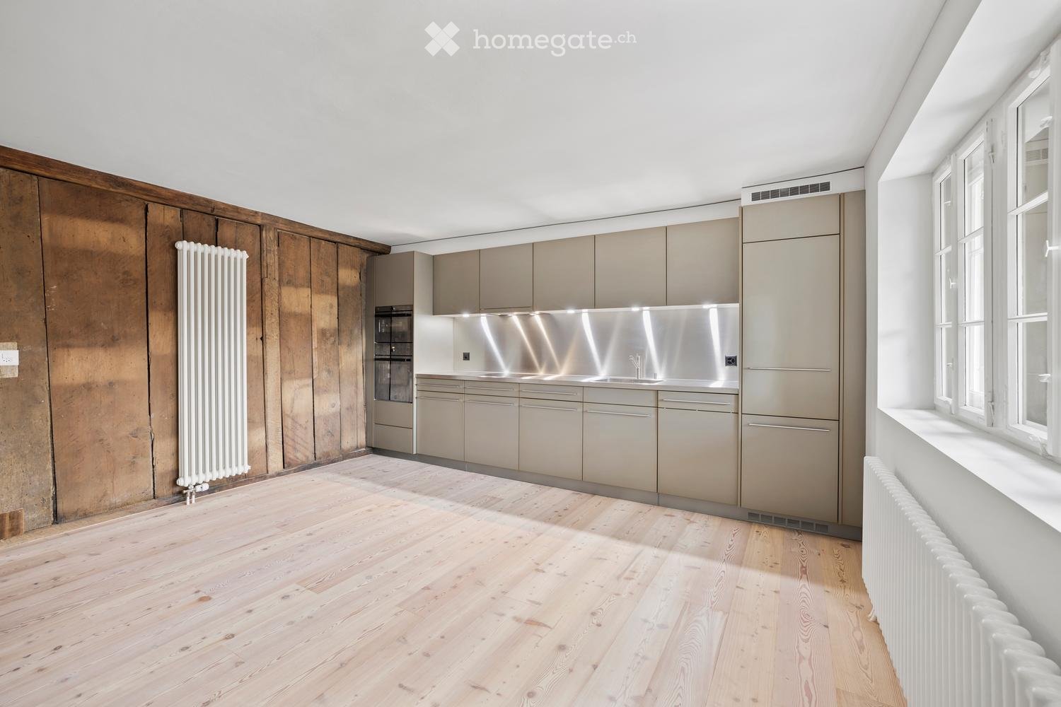 Modern kitchen with light wood flooring, stainless steel appliances, and grey cabinets. Features a wooden accent wall and large windows.