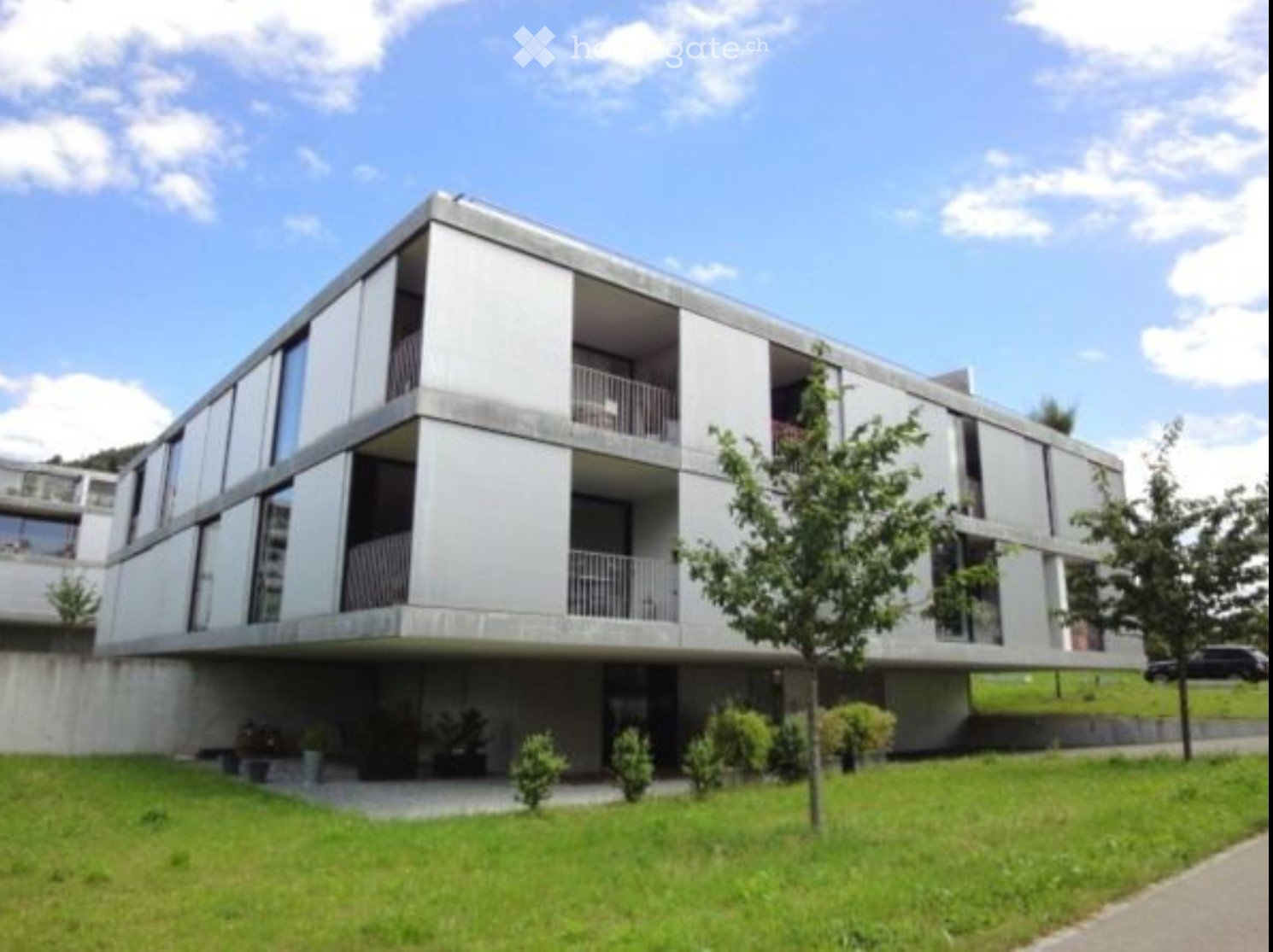 Modern, multi-story building with balconies and large windows, surrounded by a well-maintained lawn and small trees.