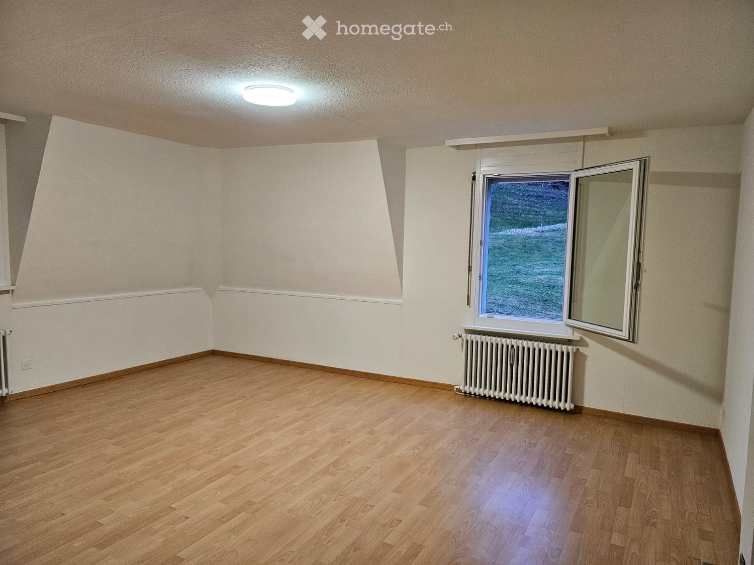 Empty room with wooden flooring, white walls, a window with a view, and a radiator under the window.