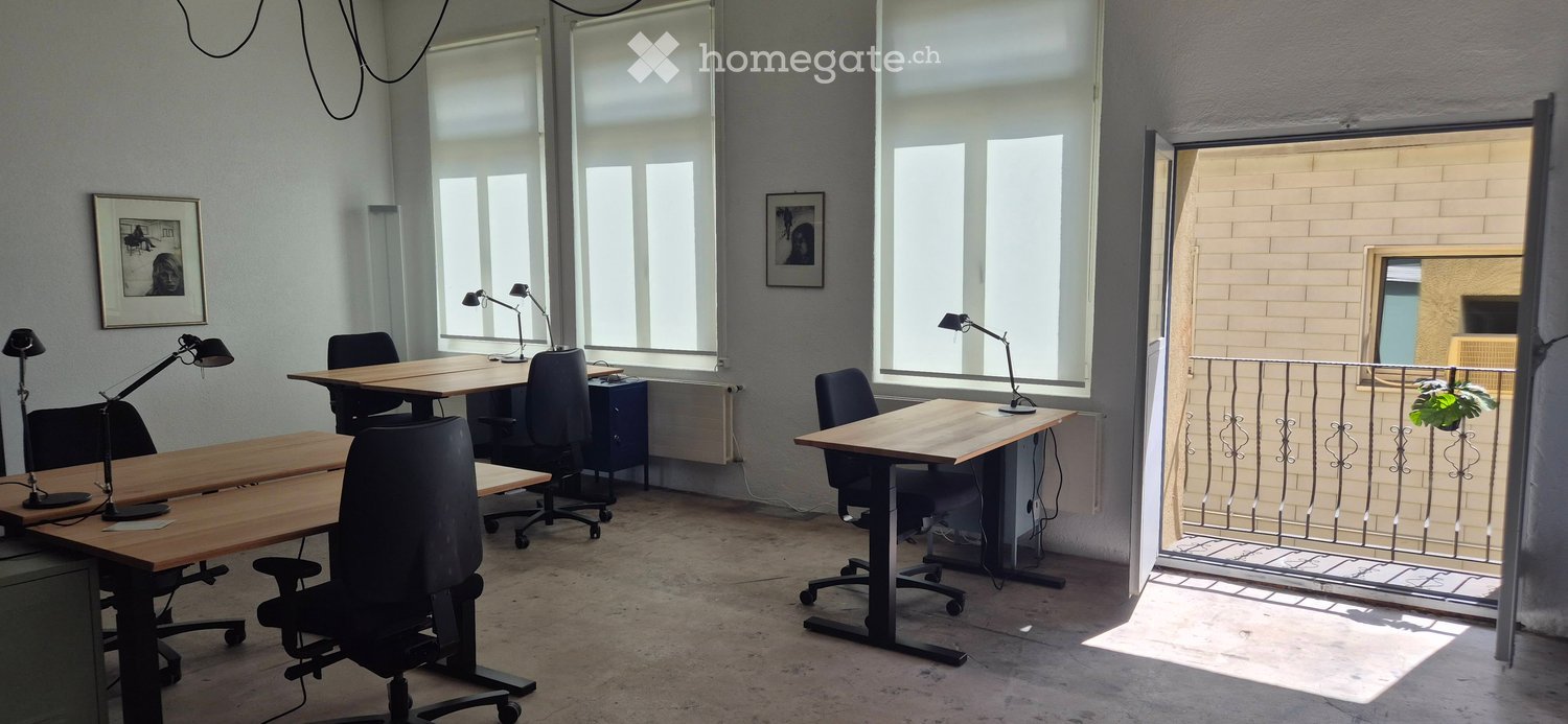 Bright, open-plan office space with large windows, wooden tables, black office chairs, adjustable lamps, and framed artwork on the walls. A balcony with a metal railing is visible through an open doorway.