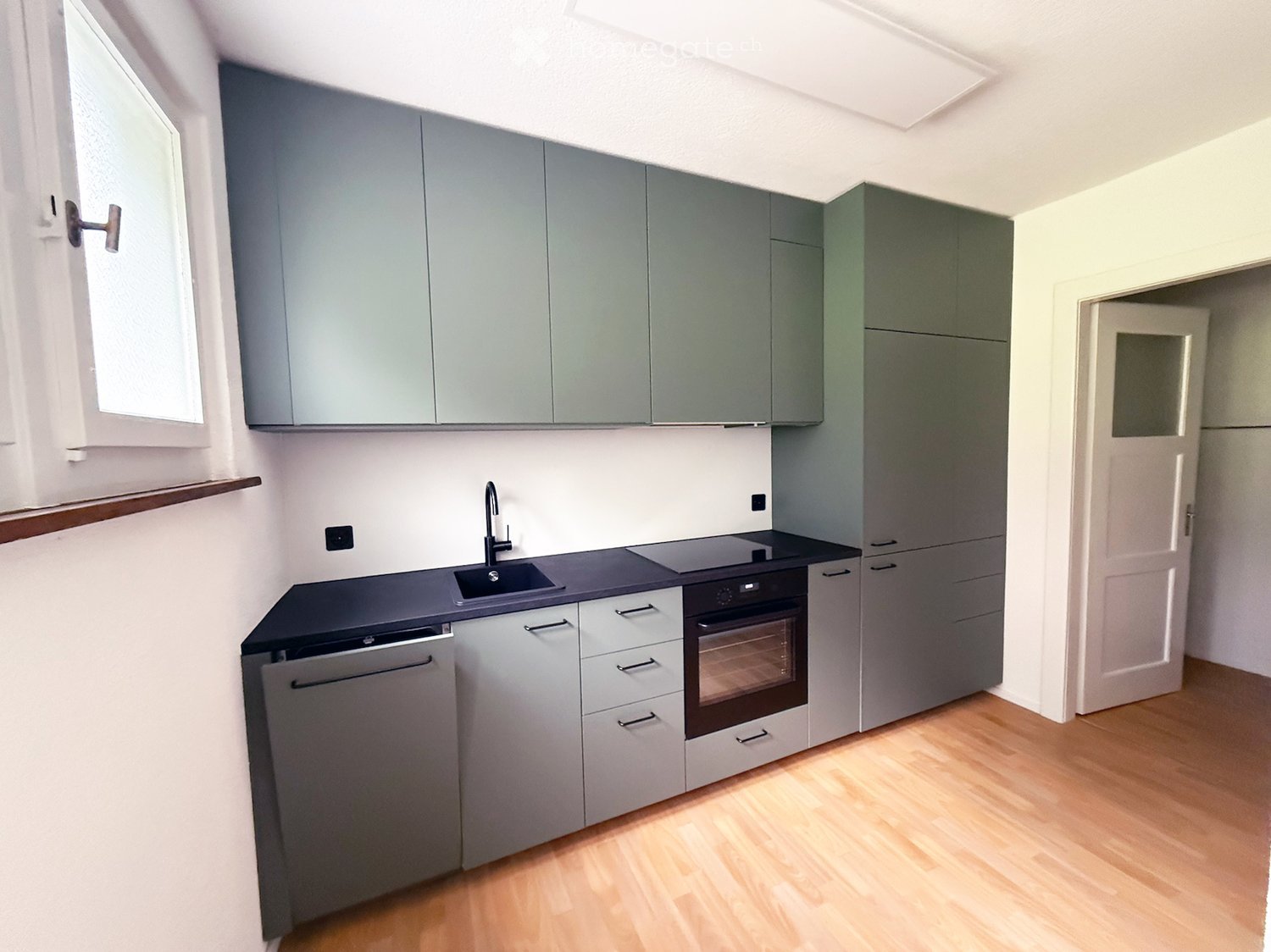 Modern kitchen with grey cabinets, black countertop, stainless steel sink, faucet, oven, and dishwasher. Light wooden flooring and white walls.