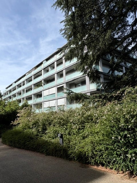 Modern multi-story apartment building with light blue facade, multiple balconies, surrounded by greenery and trees.