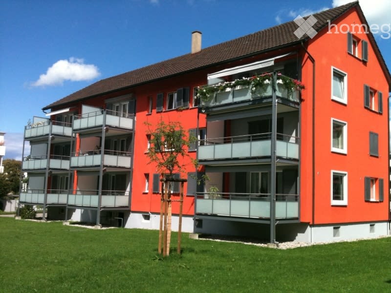 A modern, three-story apartment building with red-orange walls, multiple balconies, and a well-maintained lawn in front.