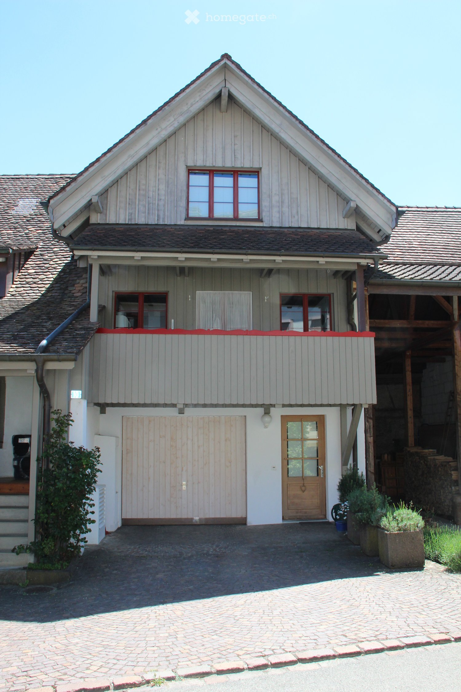 Two-story house with a gabled roof, wooden siding, and red-framed windows. Features a covered balcony area, a garage, and a paved driveway.