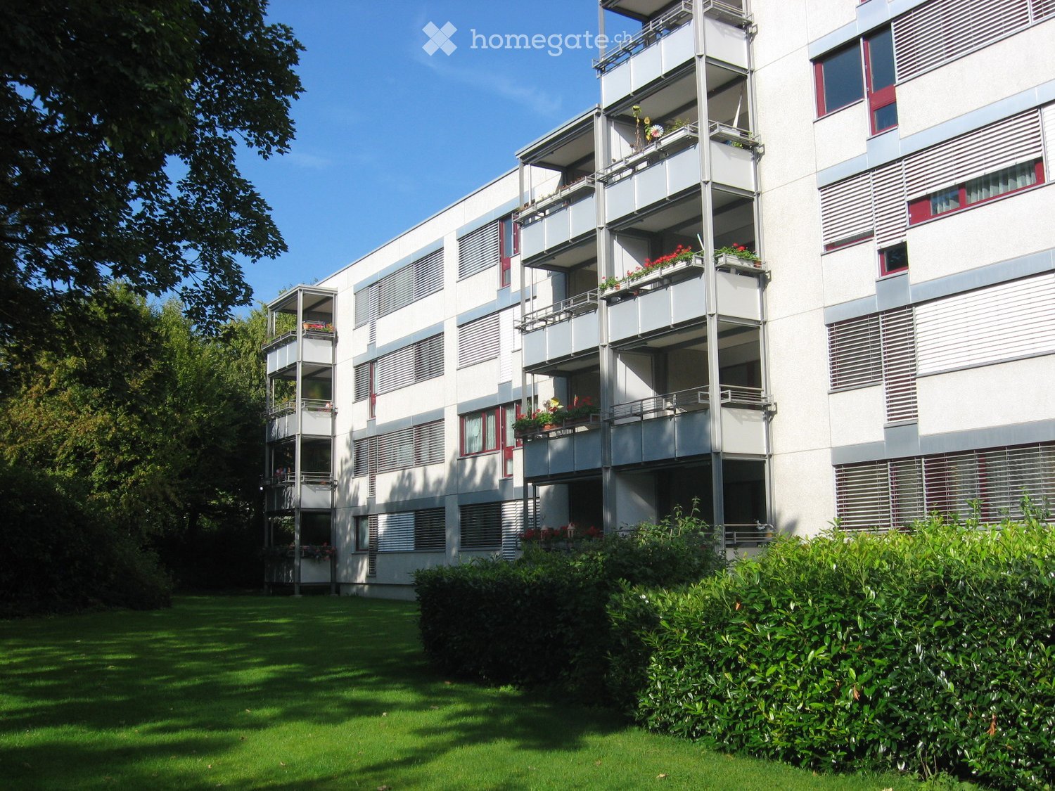 Modern multi-story apartment building with balconies, red-framed windows, and surrounding greenery.