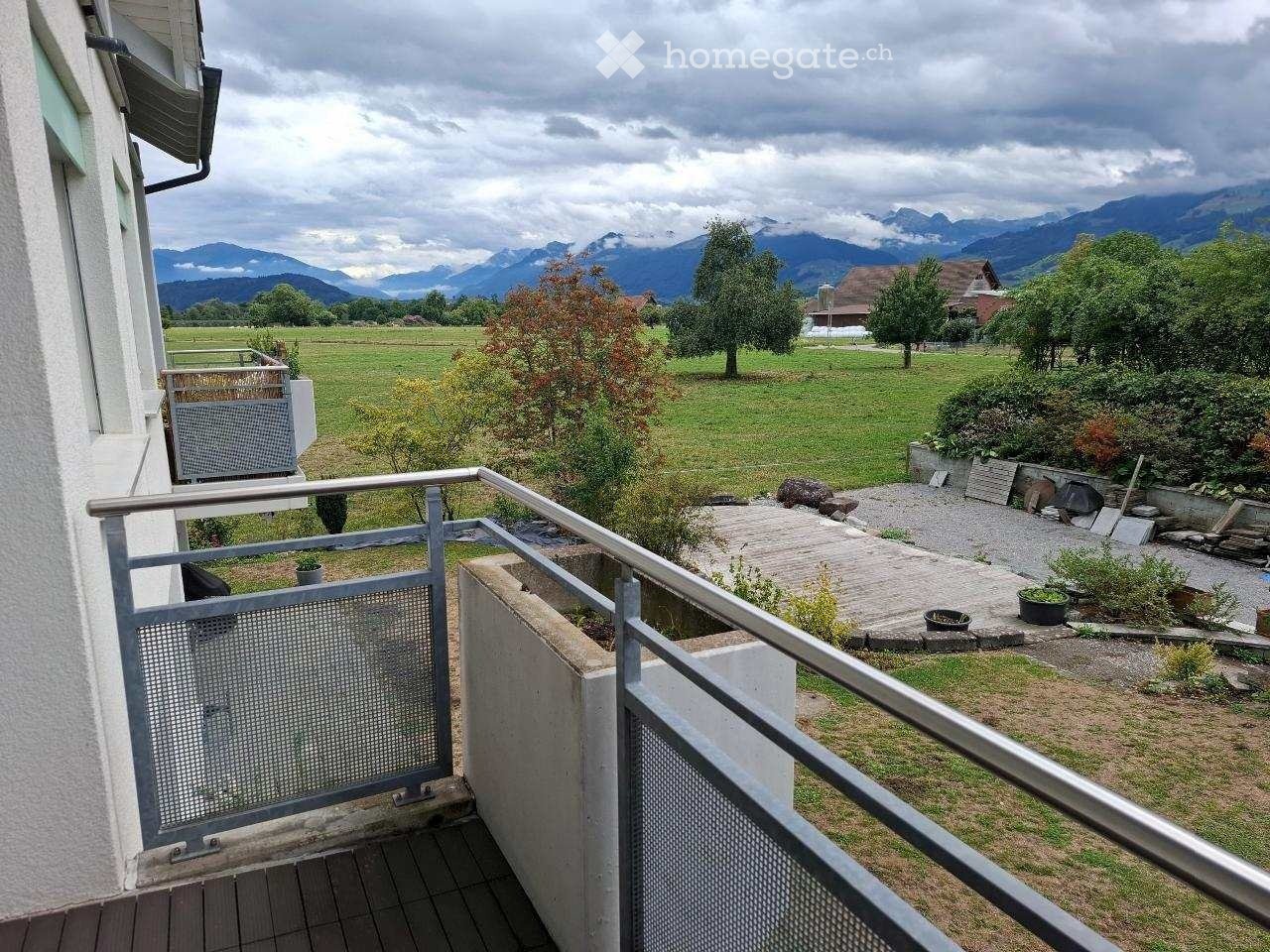 Balcony with metal railing overlooking a green field and distant mountains, with a small garden area below.