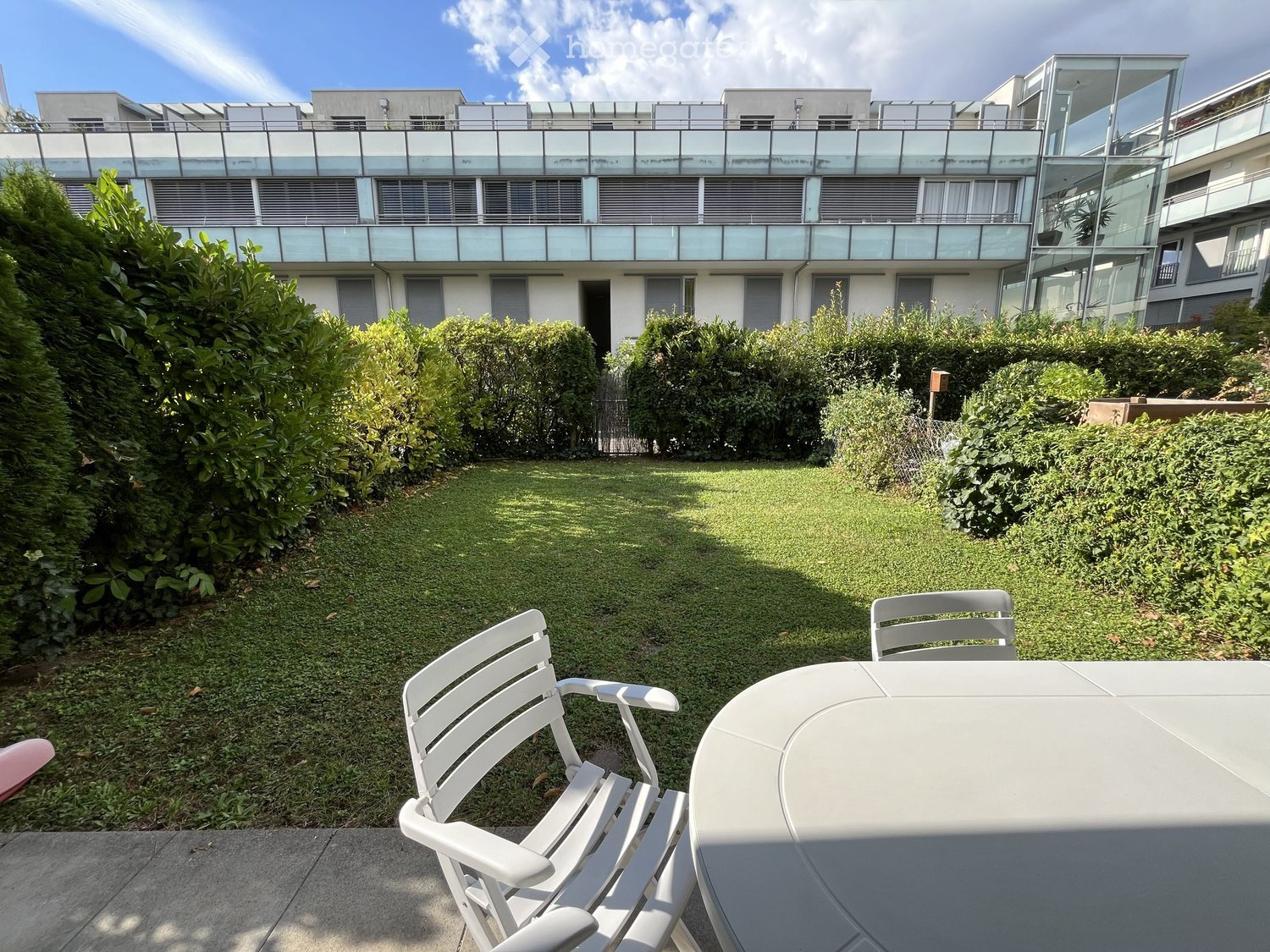 Garden area with a lawn, white plastic chairs, and a table, surrounded by bushes and a modern building with balconies.