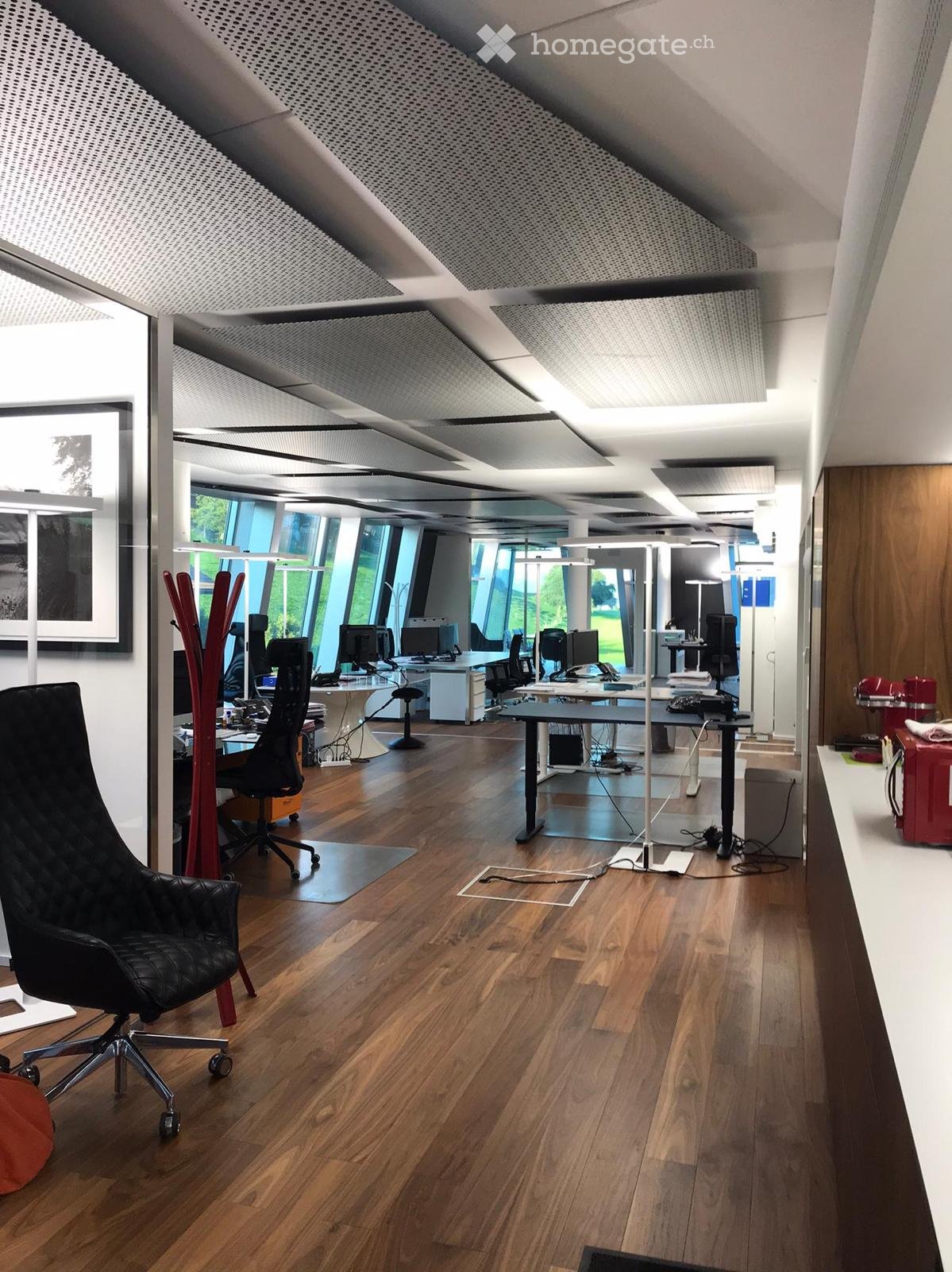 Modern office space with wooden flooring, large windows, multiple desks with computers, chairs, and a stylish black leather chair in the foreground.