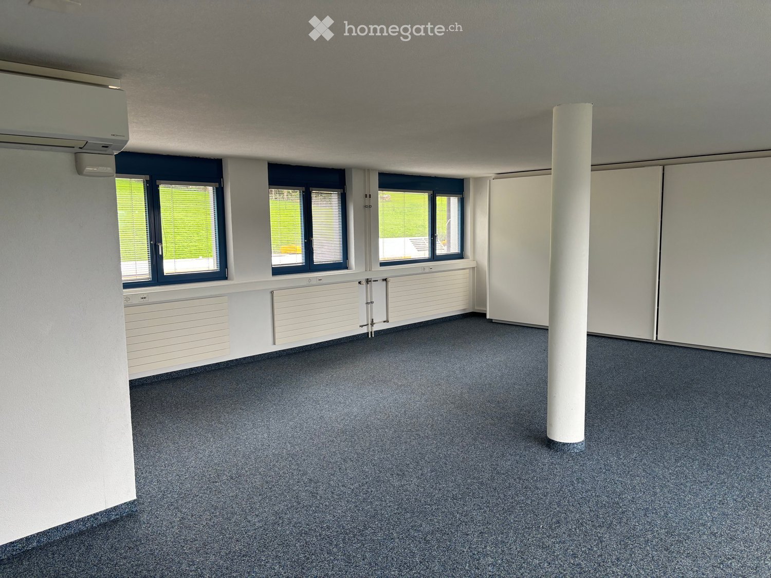 Empty room with grey carpeted floor, white walls, large windows with blinds, and a central white pillar.