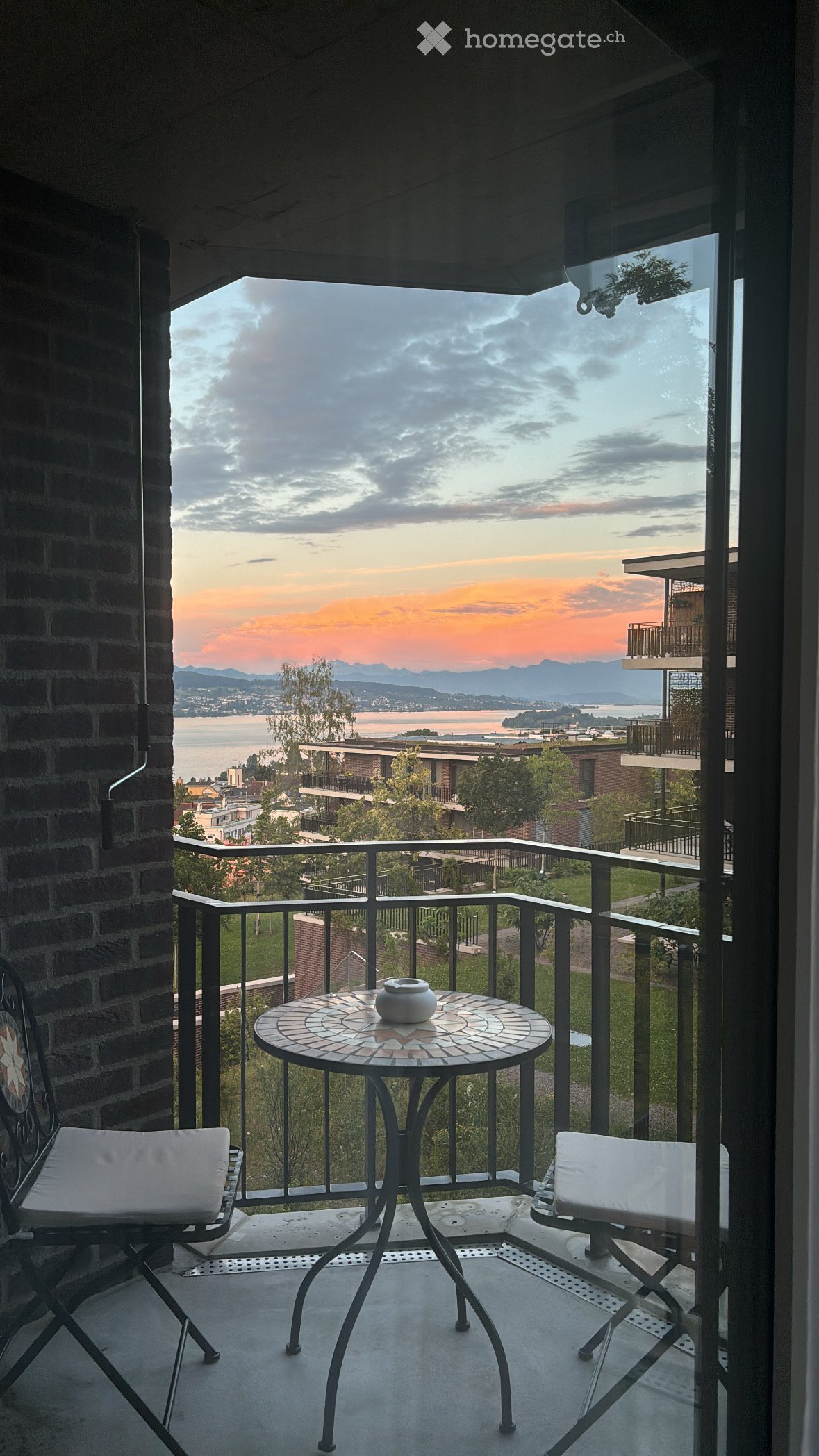 Balcony with a round mosaic table, two white cushioned chairs, and a decorative vase, overlooking a scenic view of a lake and distant mountains.