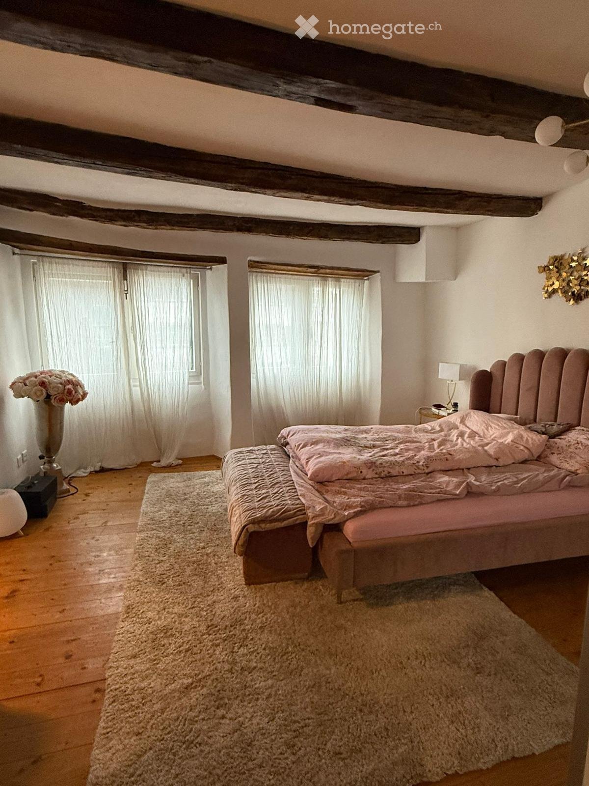 Cozy bedroom with wooden floors, exposed wooden beams, large windows with white curtains, a plush bed with pink bedding, a nightstand with a lamp, a vase with flowers, and a soft white rug.