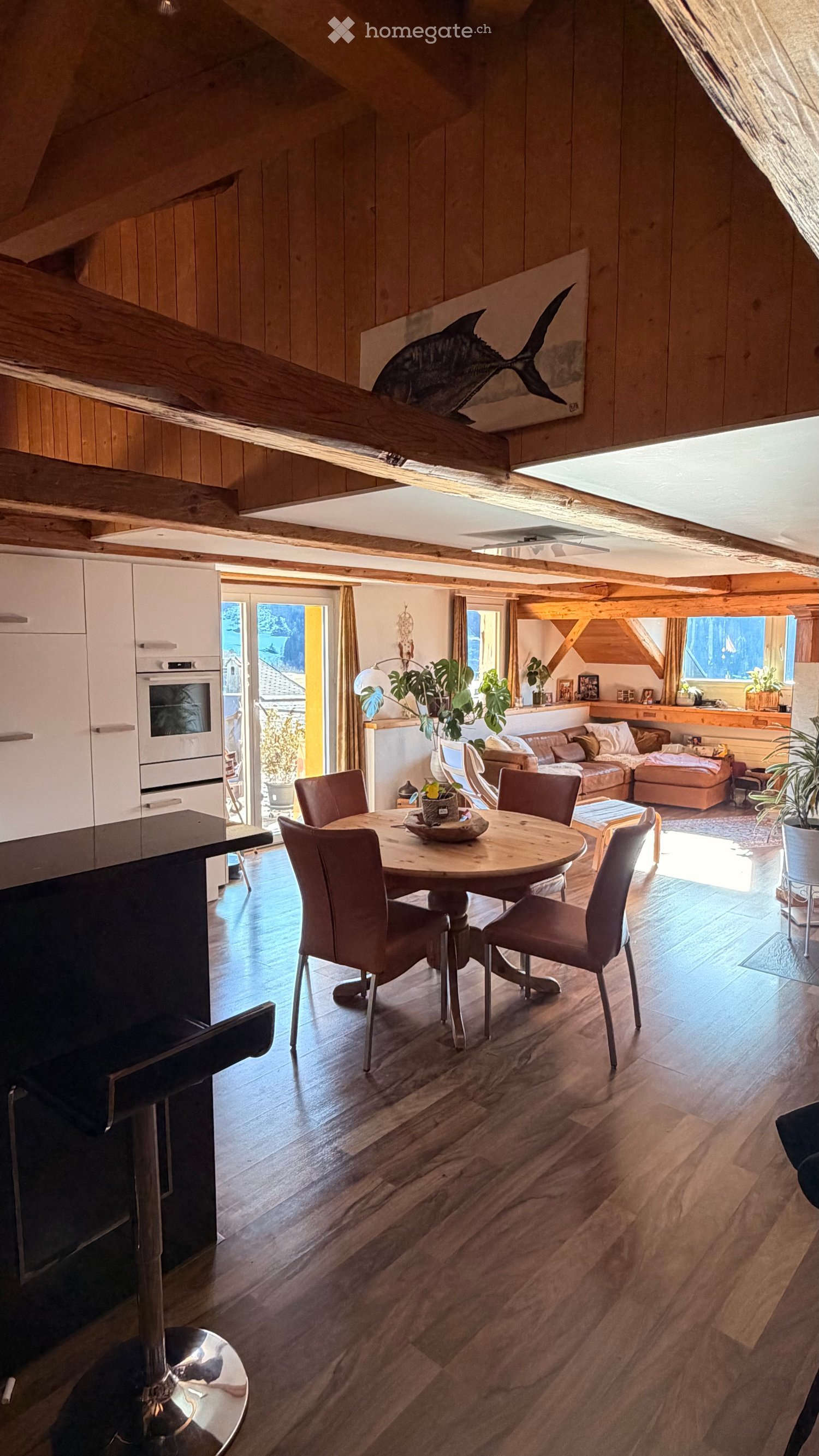 Open-plan living and dining area with wooden floors, exposed wooden beams, a round dining table with six chairs, a sofa with an ottoman, kitchen cabinets, and a modern oven. Large windows and a sliding glass door lead to a balcony.