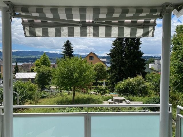 Balcony with striped awning, overlooking a green landscape with trees, houses, and distant mountains.