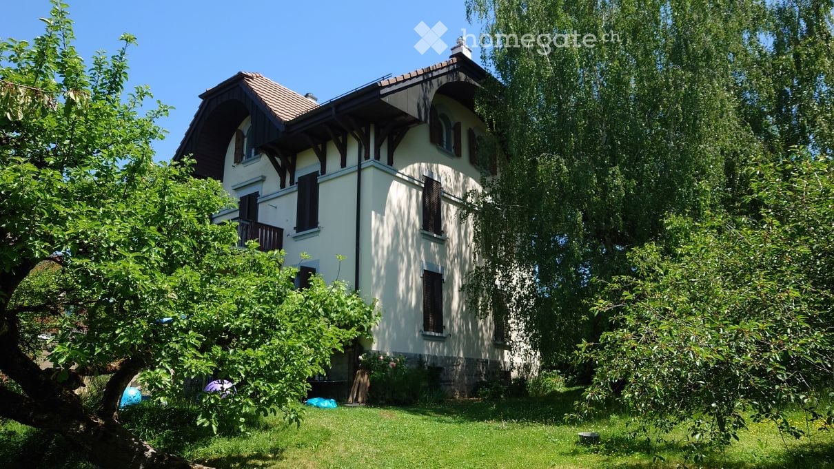 Two-story house with a gabled roof, white exterior, dark shutters, and a balcony on the upper floor, surrounded by trees and a grassy lawn.