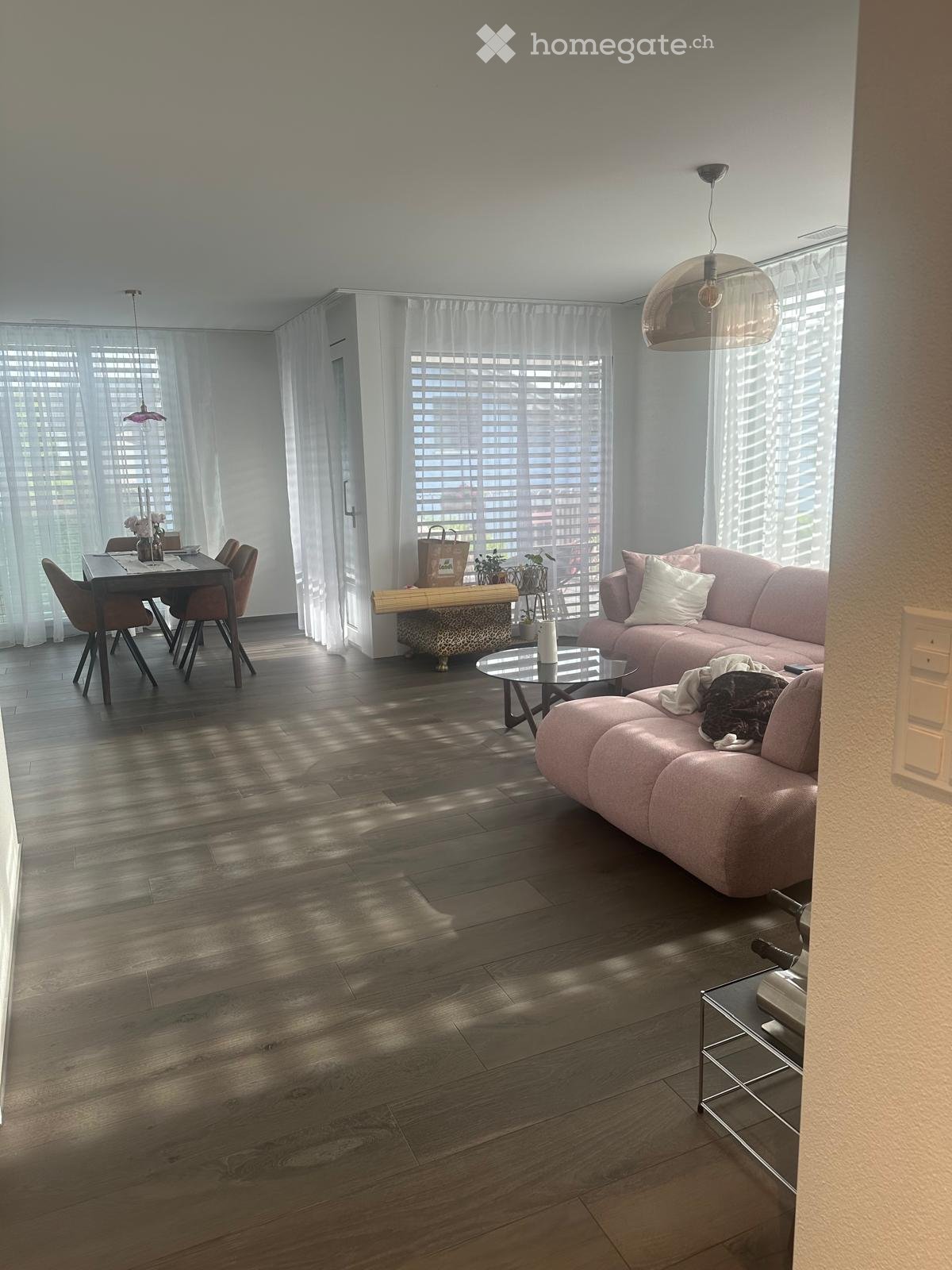 Modern living room with a pink sofa, glass coffee table, dining area, and large windows with blinds.