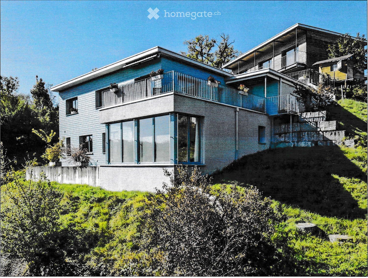 Modern two-story house with blue and grey exterior, large windows, balconies, and built on a hillside with surrounding greenery.