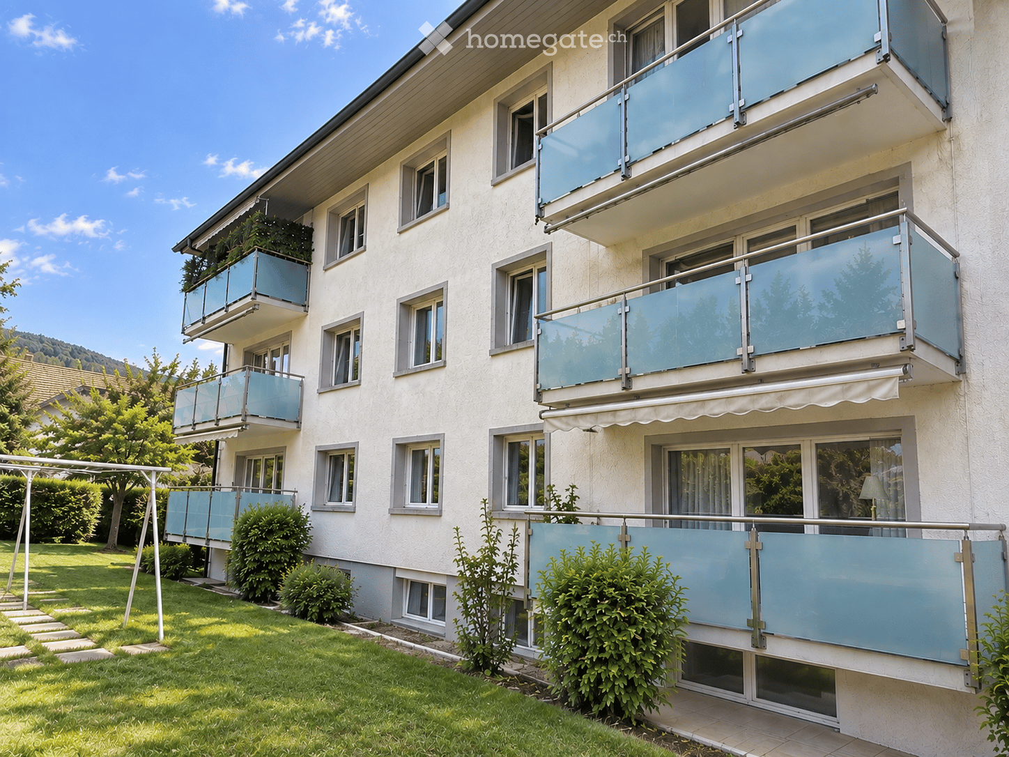 Multi-story residential building with light-colored facade, multiple windows, and glass-enclosed balconies. Green lawn and shrubs in front.