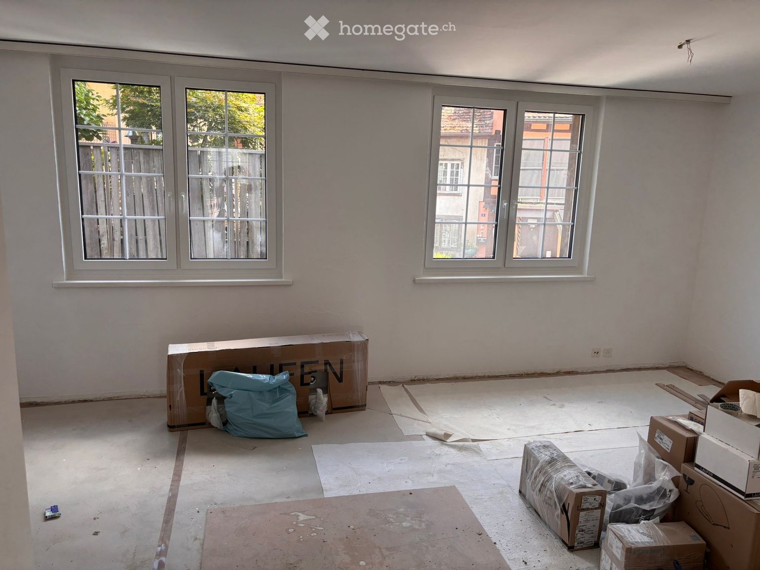 Empty room with white walls, large windows, and scattered boxes and bags on the floor.